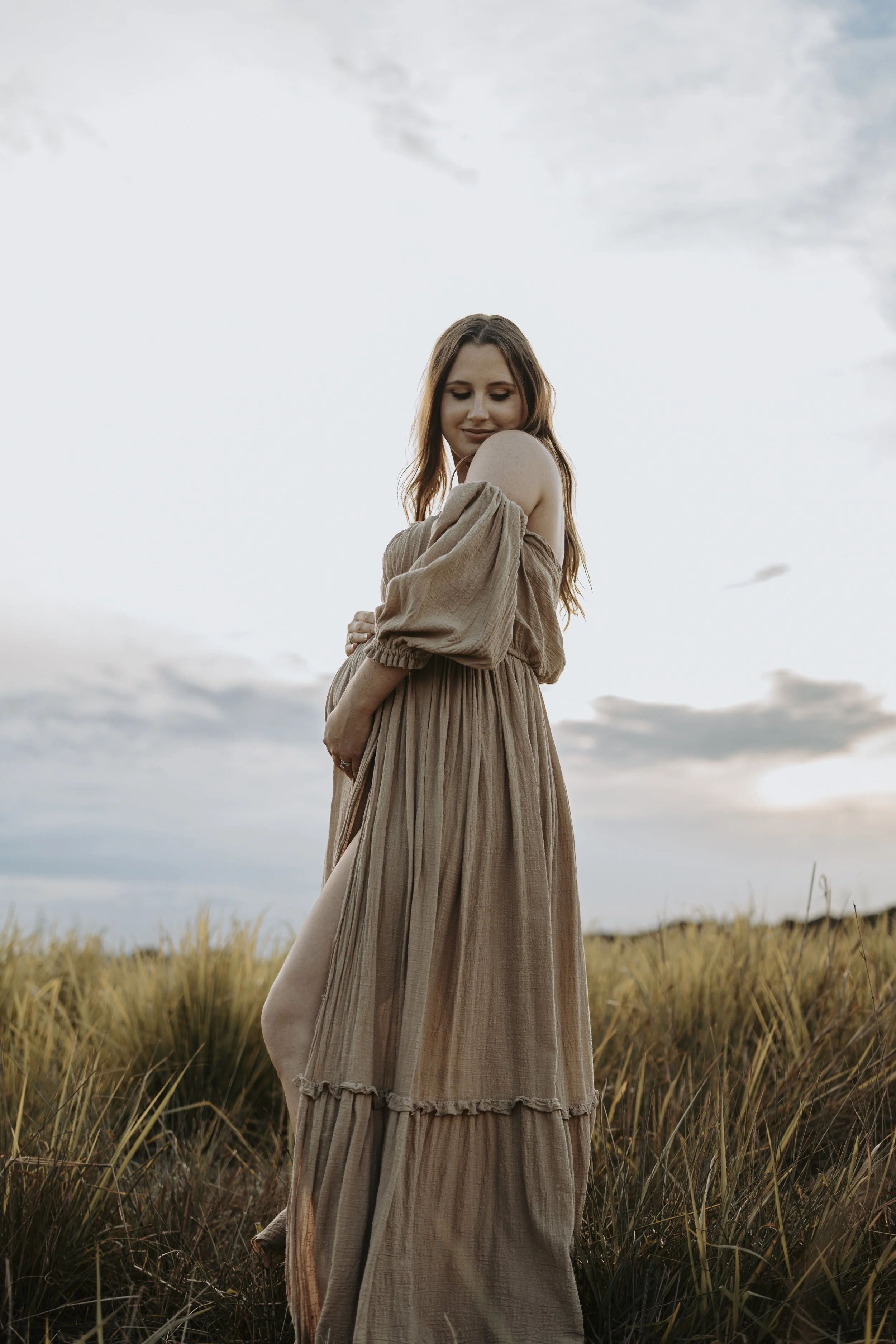 A woman in a beige off-shoulder dress standing in a field of tall grass during sunset.