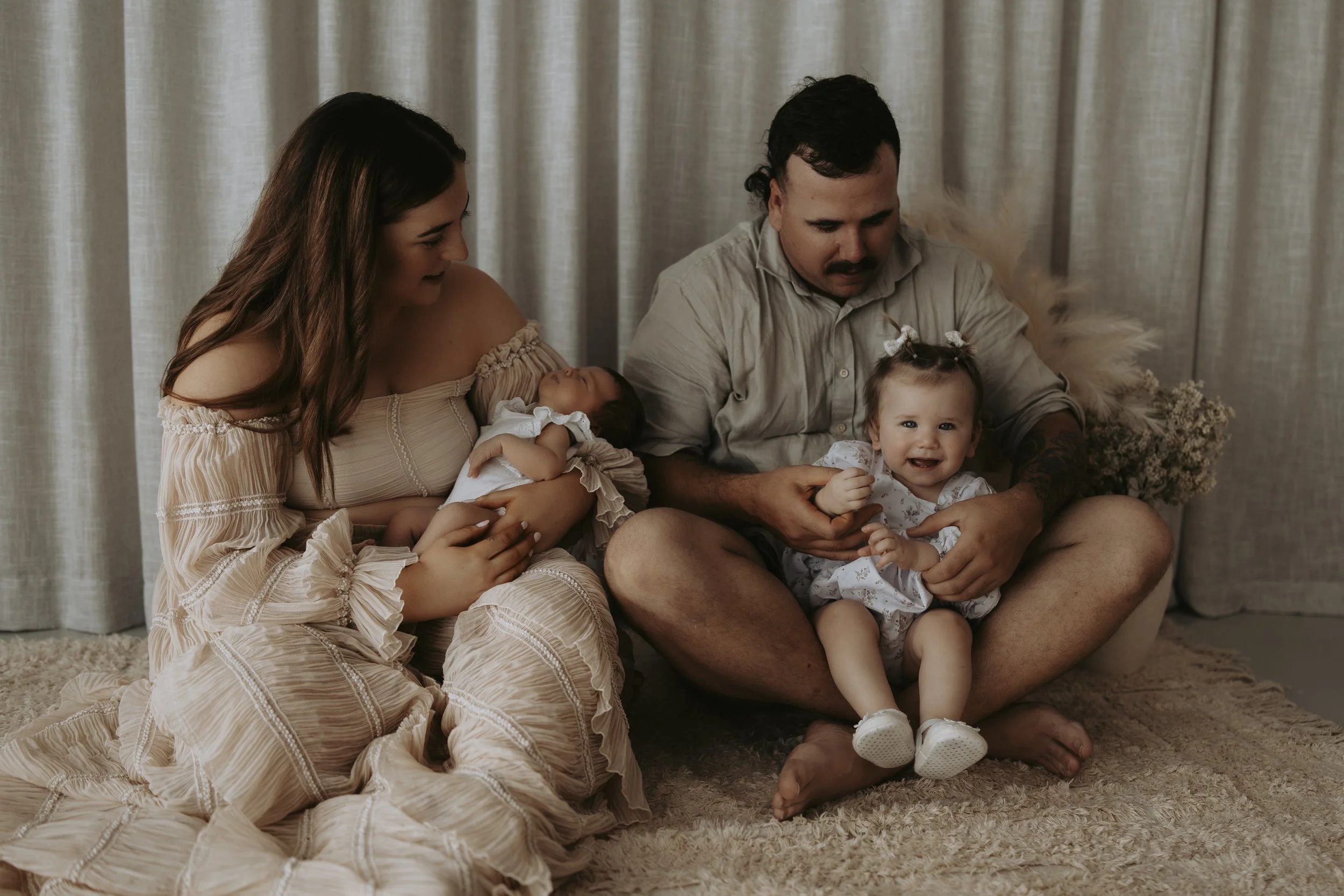 Family scene with mother, father, and two young children sitting on a soft carpet in front of beige curtains.