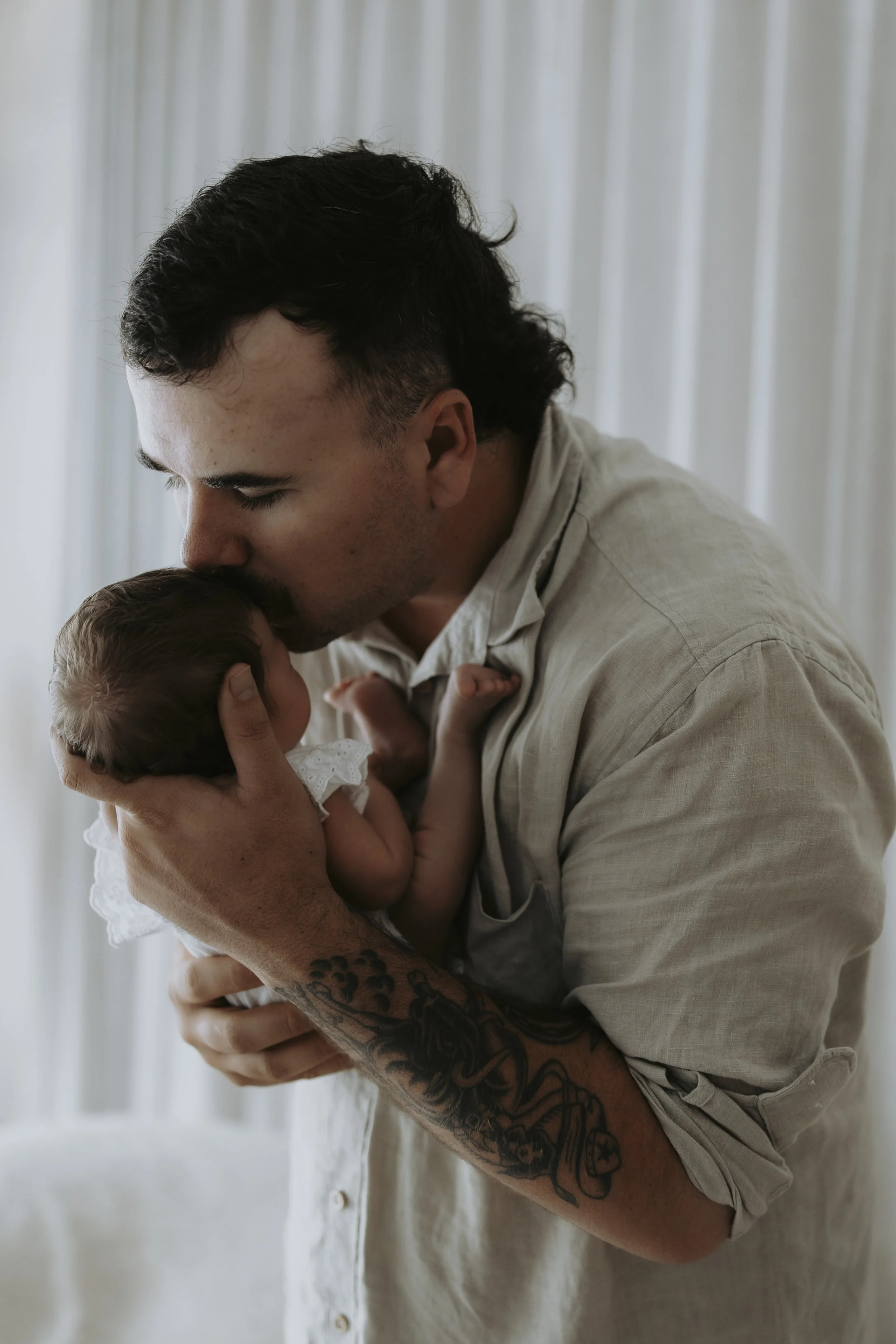 A man with dark hair and tattoos on his arm holds a newborn baby close, kissing the baby's forehead in a tender moment.