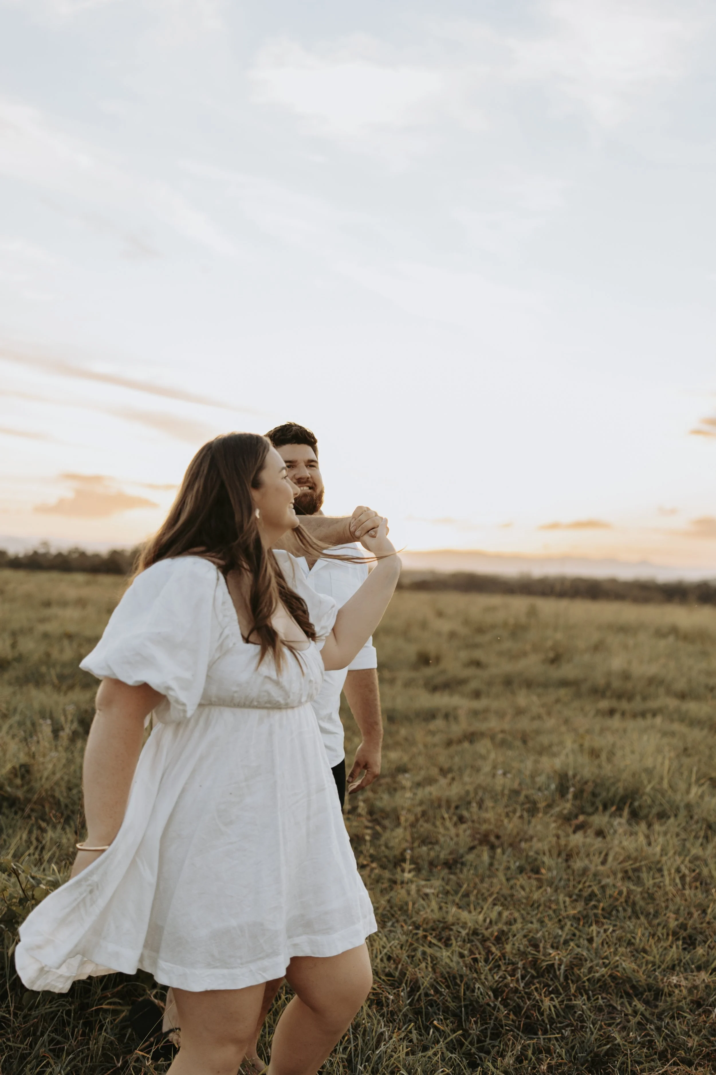 A woman and a man standing in a grassy field during sunset, with the woman looking at the woman and smiling, both dressed in white.