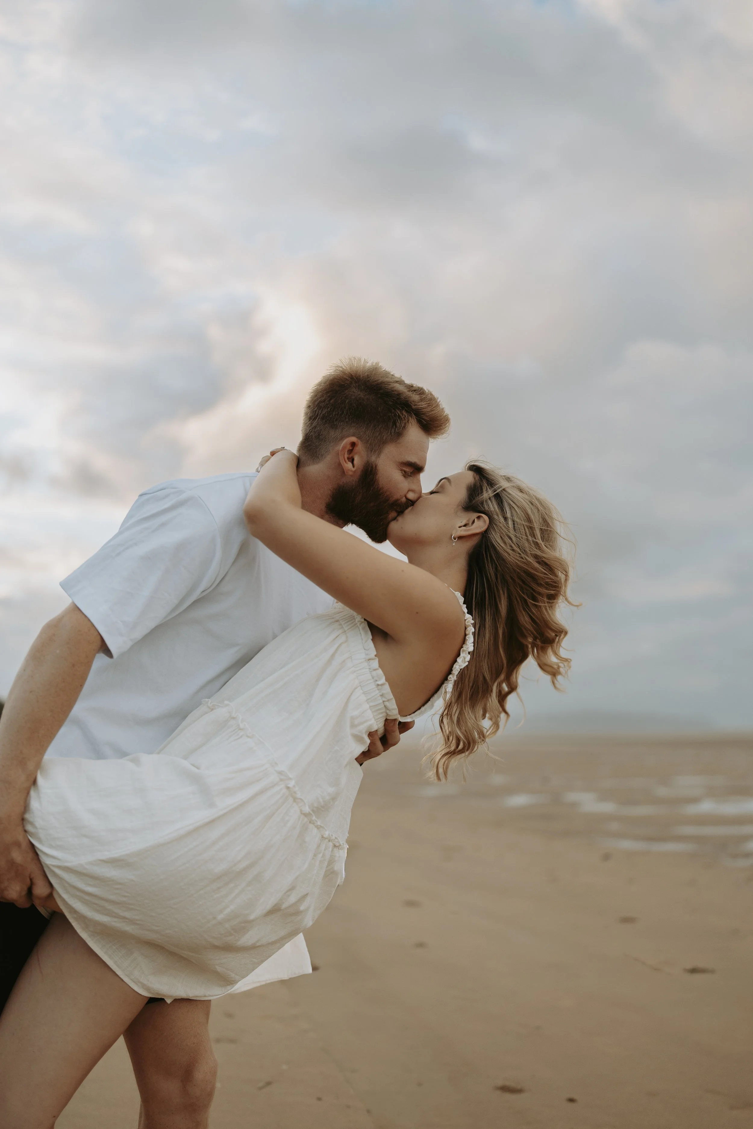 A couple kissing on the beach, with the man holding the woman as she leans back, both wearing light-colored clothing, under a cloudy sky.