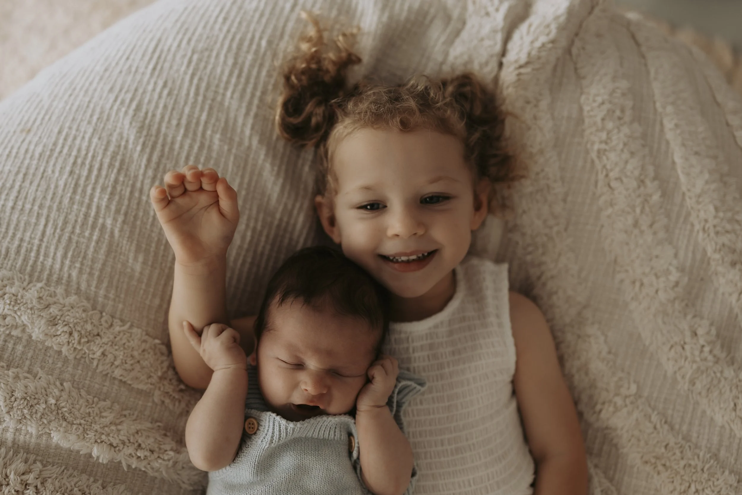 A young girl lying on a bed with an infant, both smiling and looking happy.