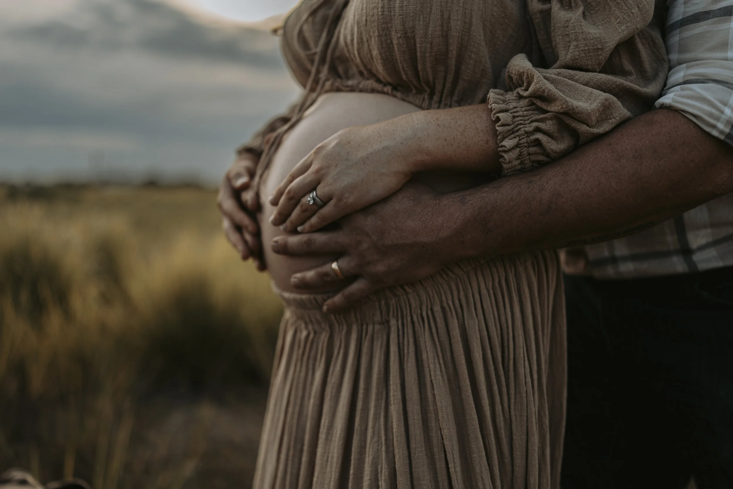 A pregnant woman and a man, both wearing rings, holding her baby bump outside in a natural setting.