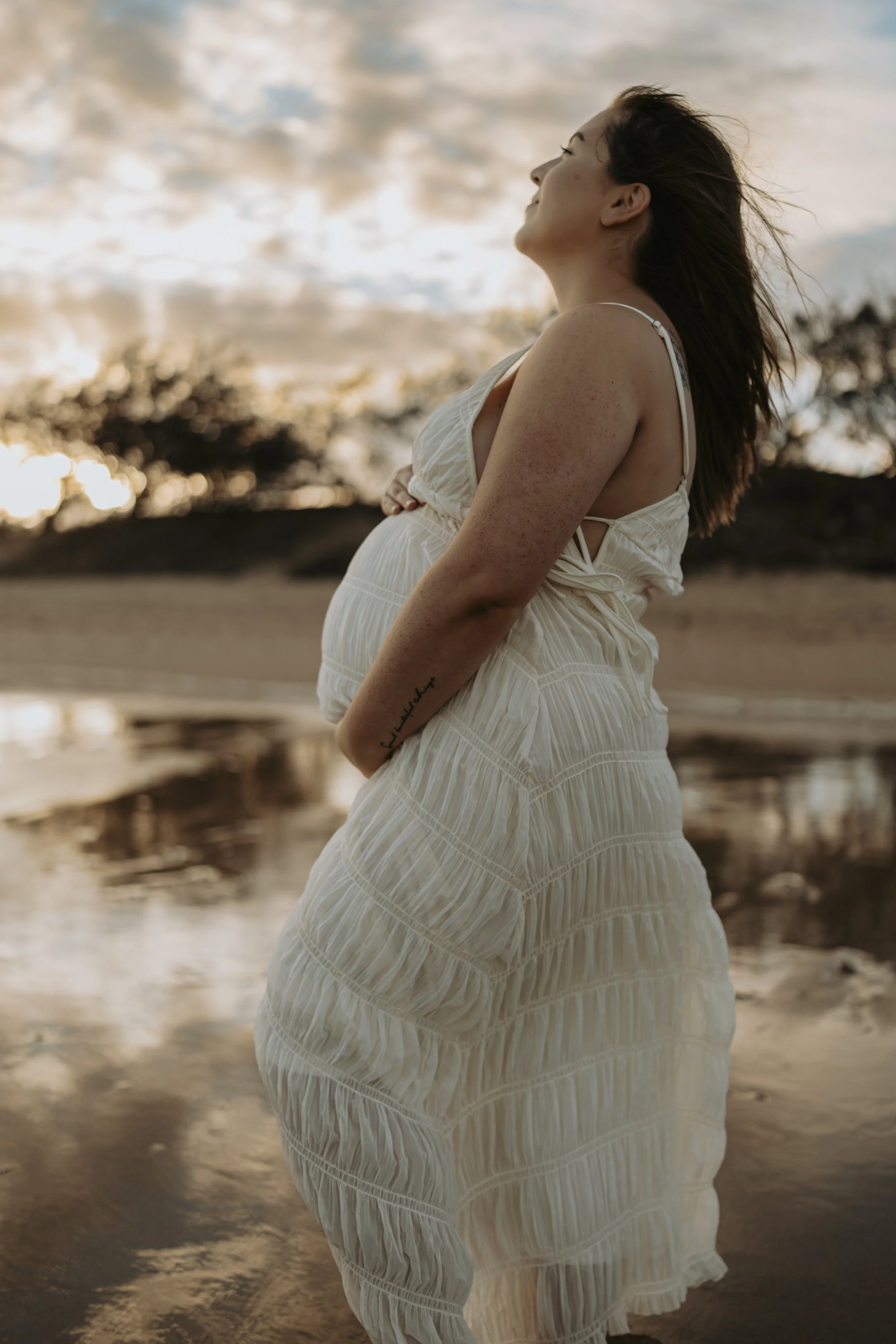 A pregnant woman in a white dress standing on a beach during sunset with her hands on her belly, looking upward with her hair blowing in the wind.
