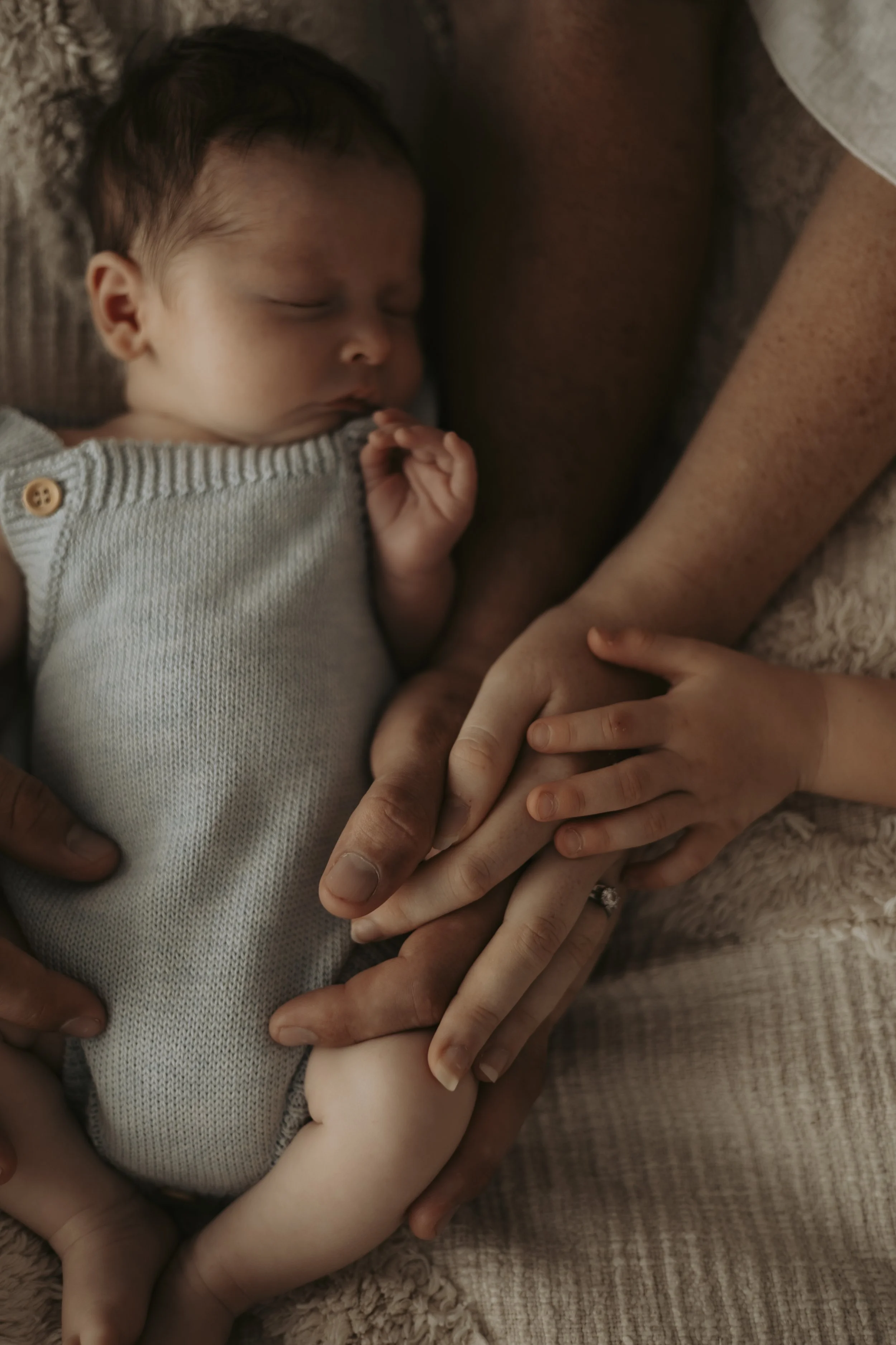 A sleeping baby lying on a soft surface while holding an adult's hand and resting its head on the adult's arm.