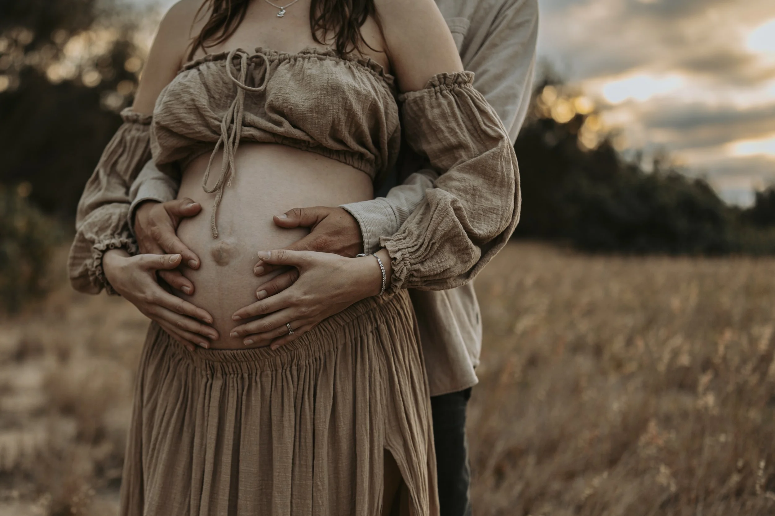 A pregnant woman with a man embracing her from behind, both with their hands on her belly, standing in a field during sunset.