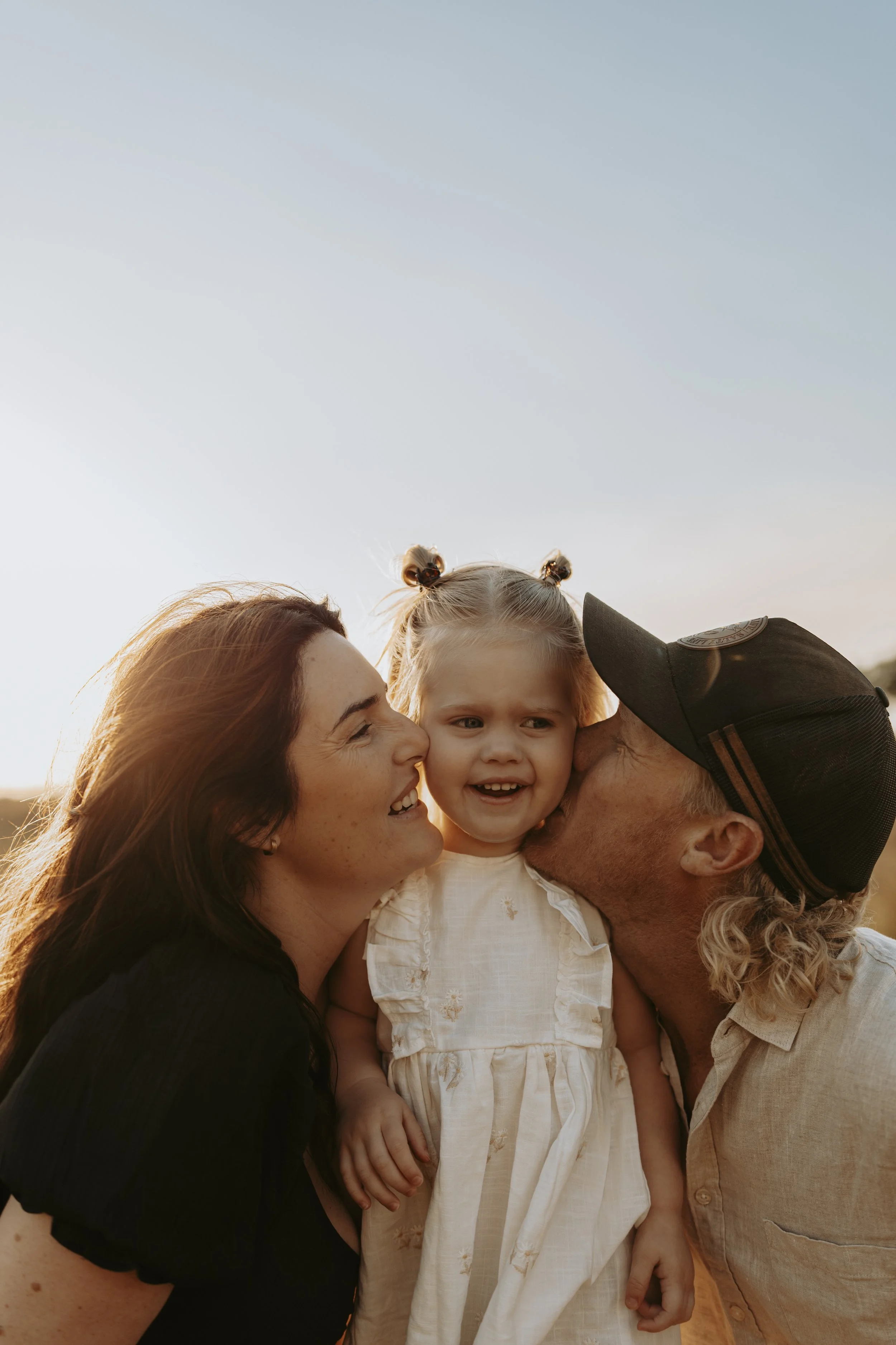 Parents kissing their young daughter on the cheeks outdoors at sunset.