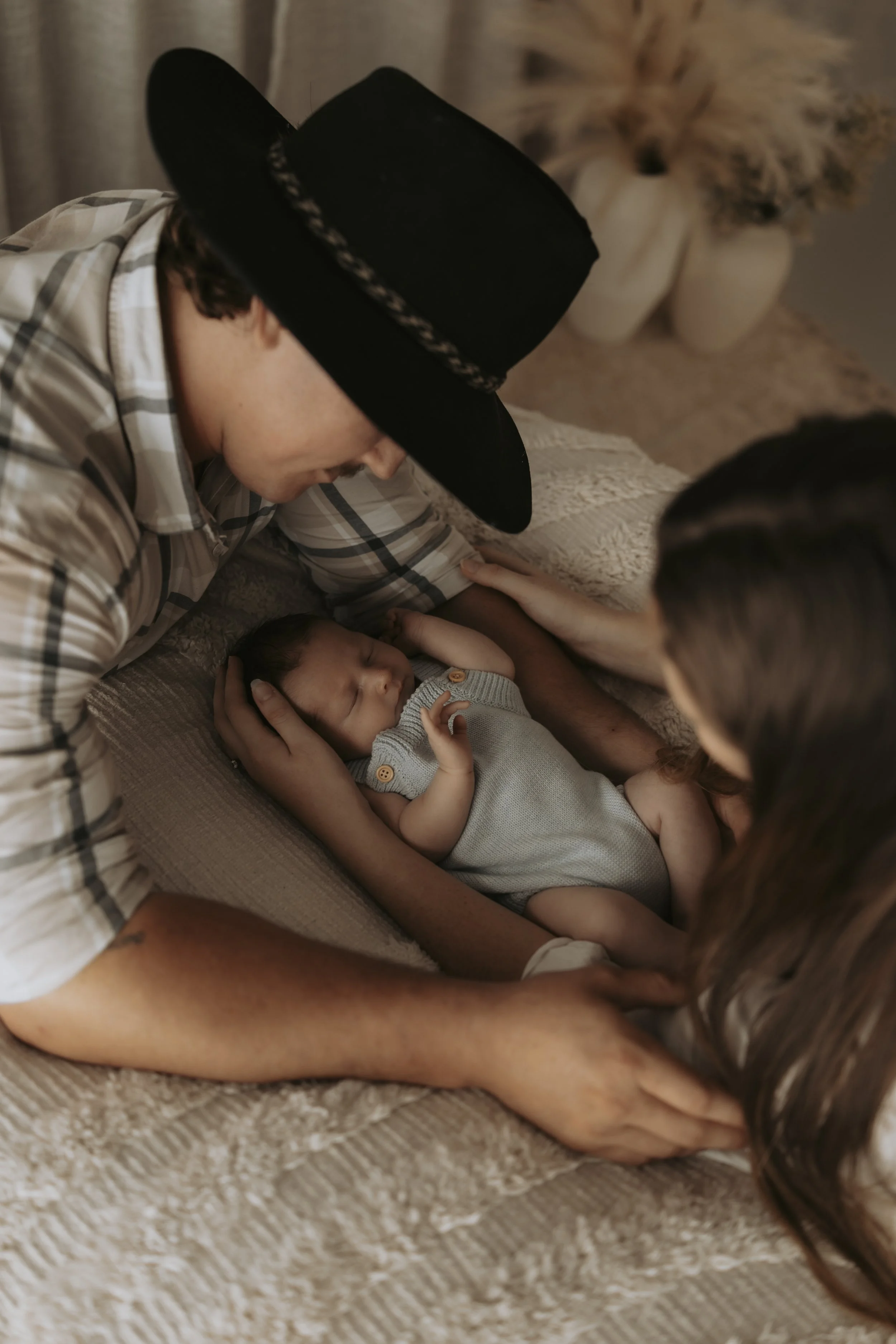A young man and woman are holding a newborn baby on a bed, with the man wearing a black hat and plaid shirt, and the woman with long brown hair. The baby is dressed in a light gray outfit and is sleeping peacefully.