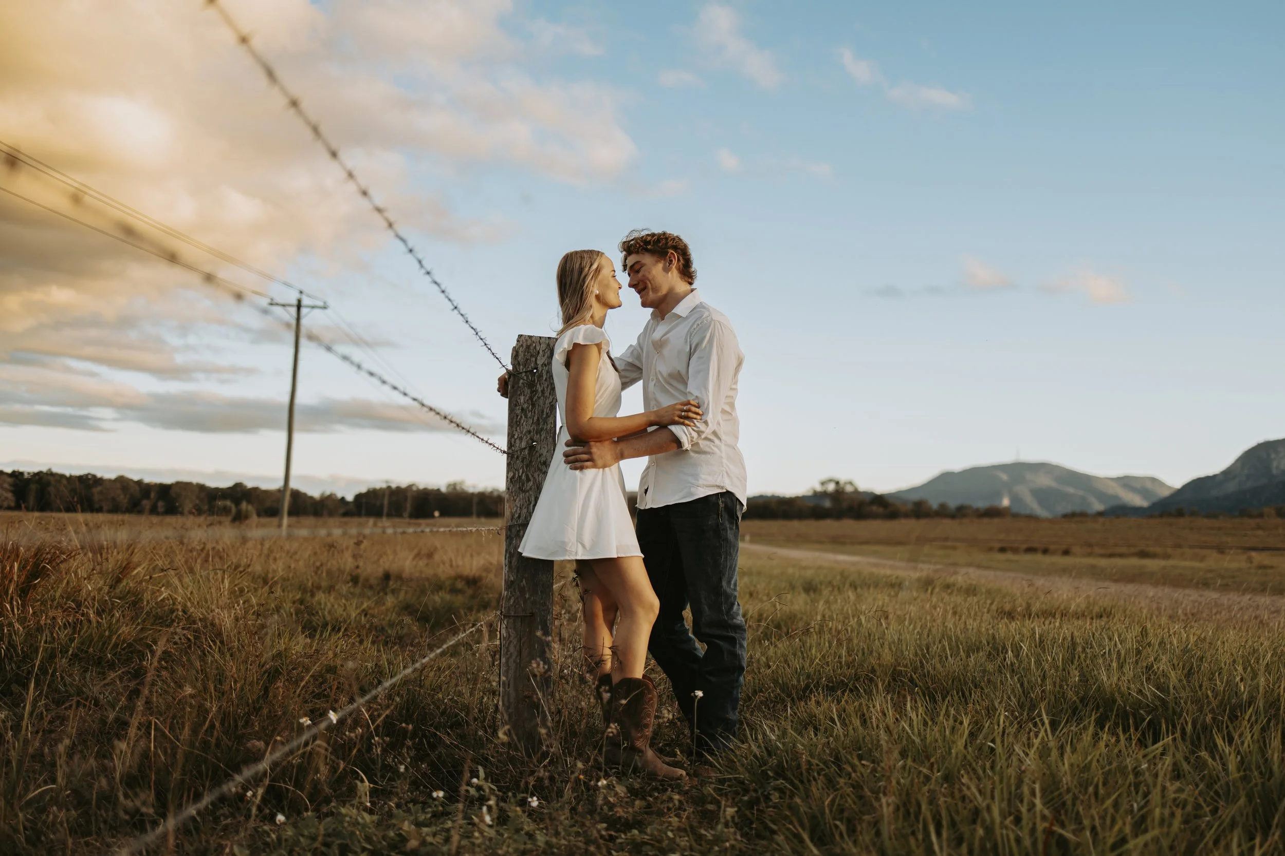 A couple stands close to each other in a field at sunset, with mountains in the background, near a barbed wire fence.
