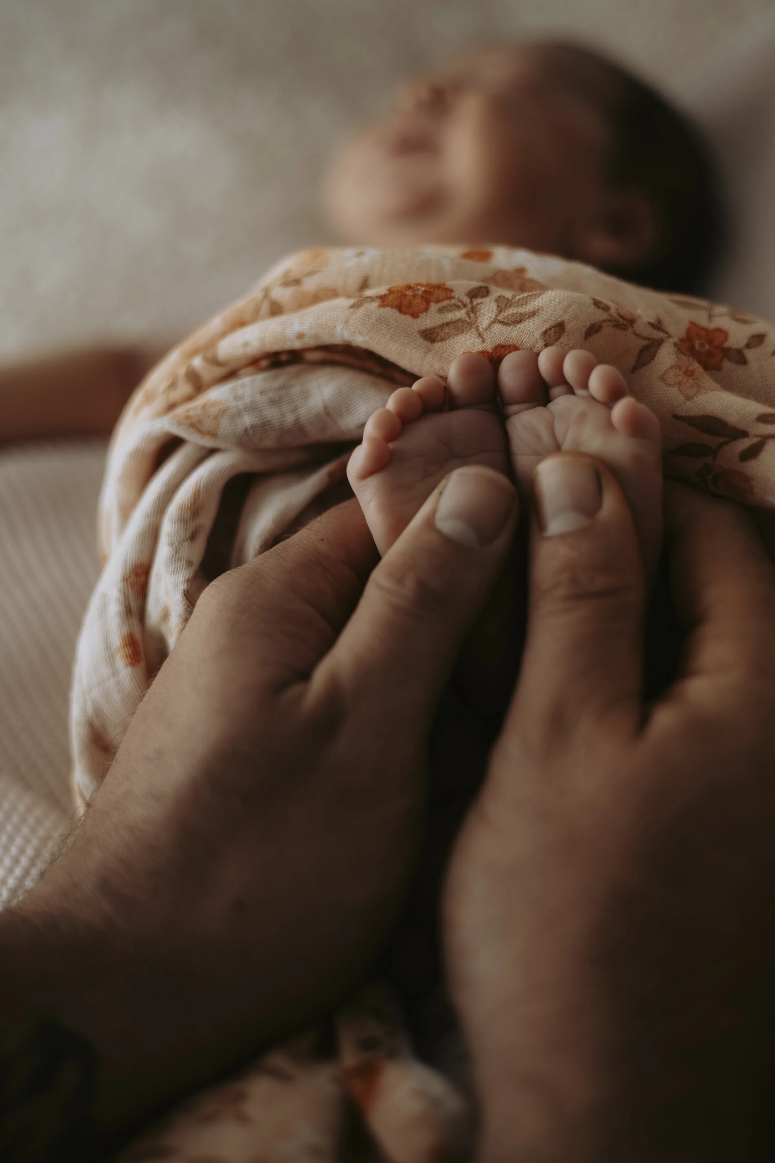 Close-up of a person's hand holding a tiny baby's foot, with the baby lying in the background. The baby is wrapped in a floral blanket.