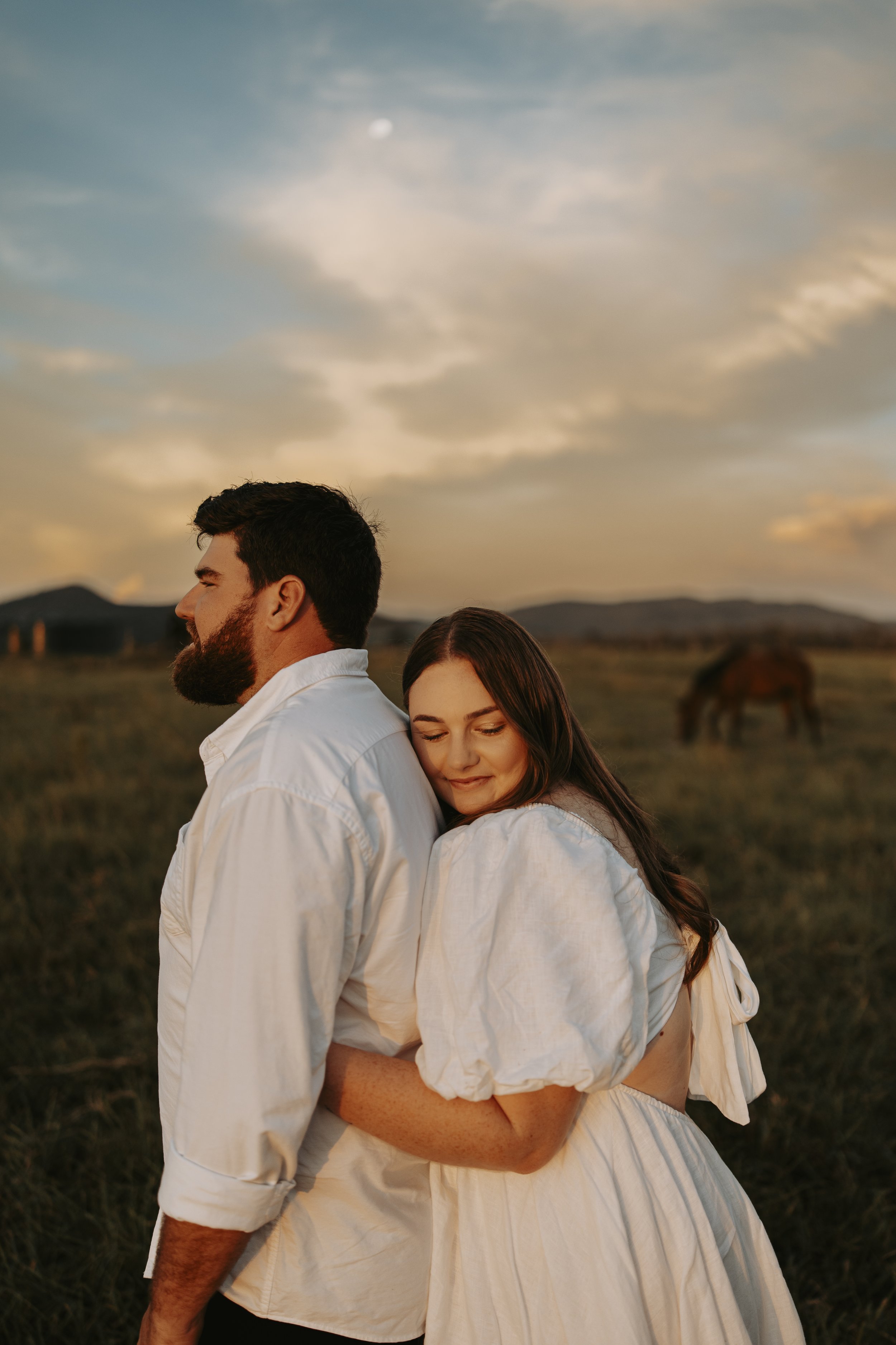 A couple stands in a field at sunset, with a horse grazing in the background, and a partly cloudy sky with the moon visible.