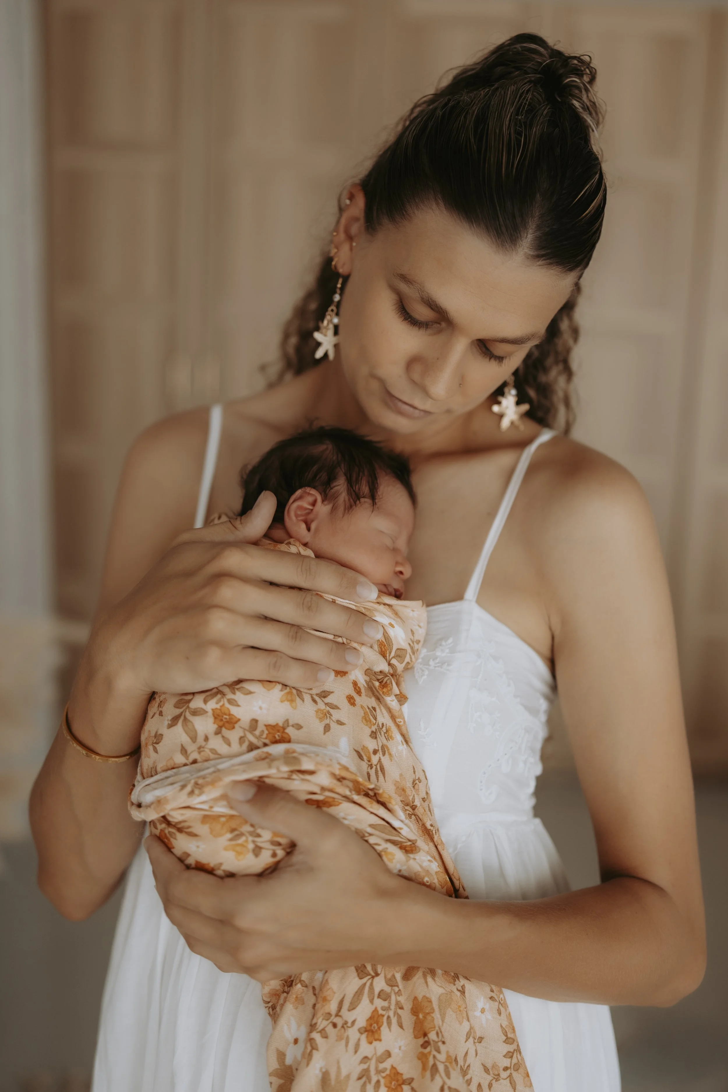 A woman in a white dress holding a sleeping newborn baby in a floral wrap, resting her head gently on the baby's head.