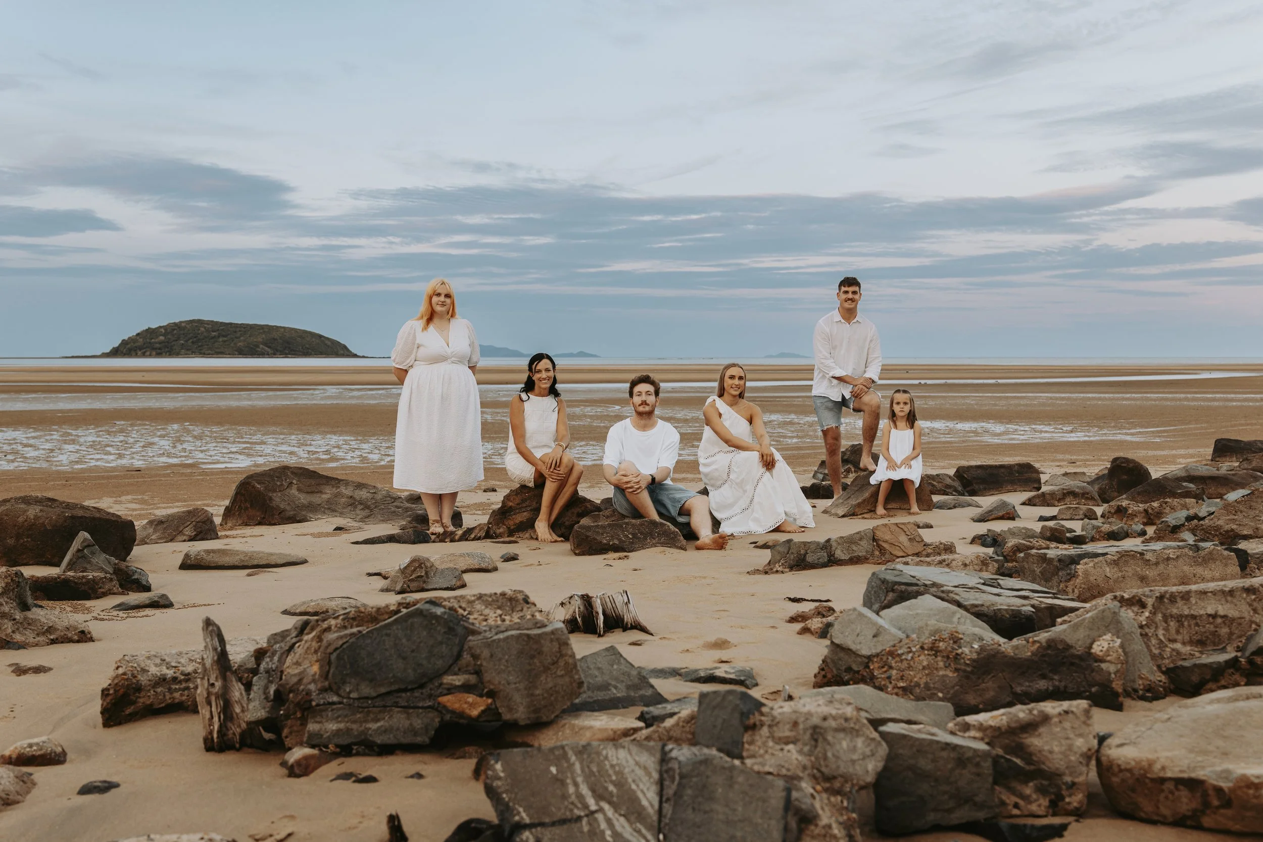 Family of seven dressed in white, sitting and standing on rocks on a sandy beach with a distant island and cloudy sky in the background.