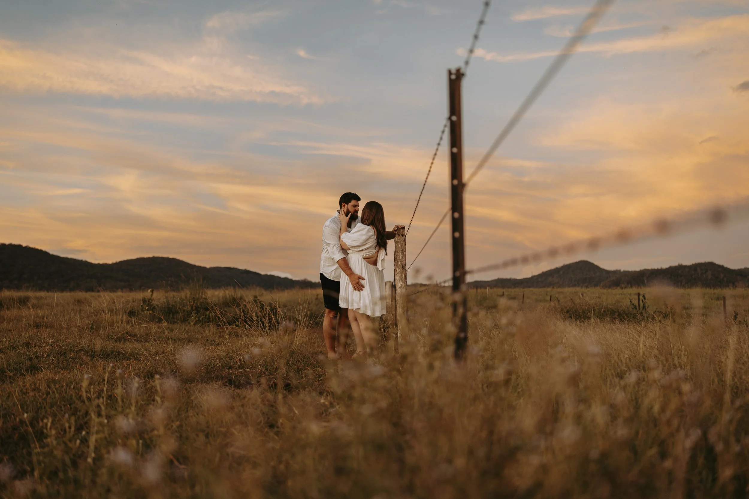 A couple standing close together in a field at sunset, near a wooden fence with mountains in the background.