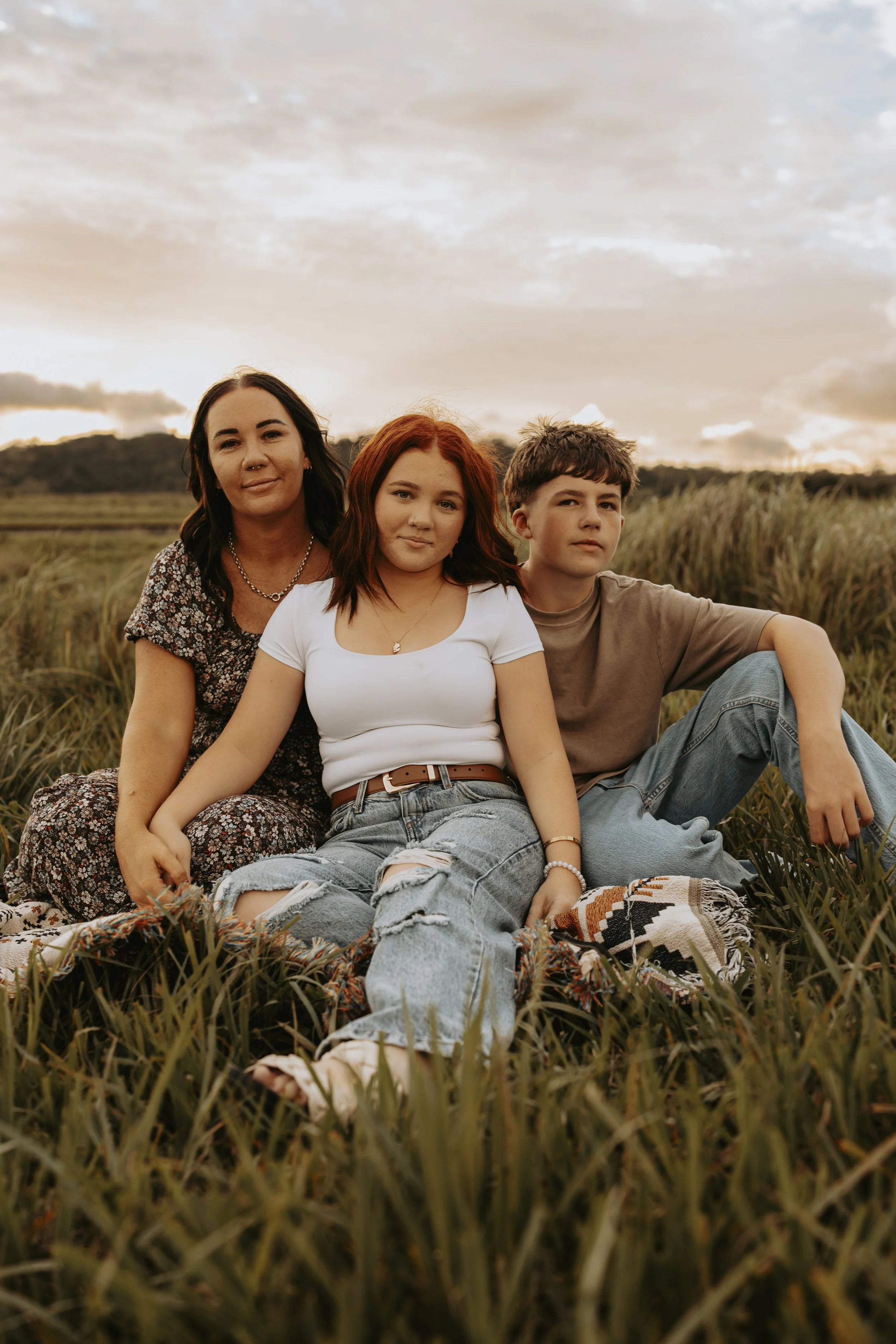 Three people sitting in a grassy field during sunset with a cloudy sky in the background.