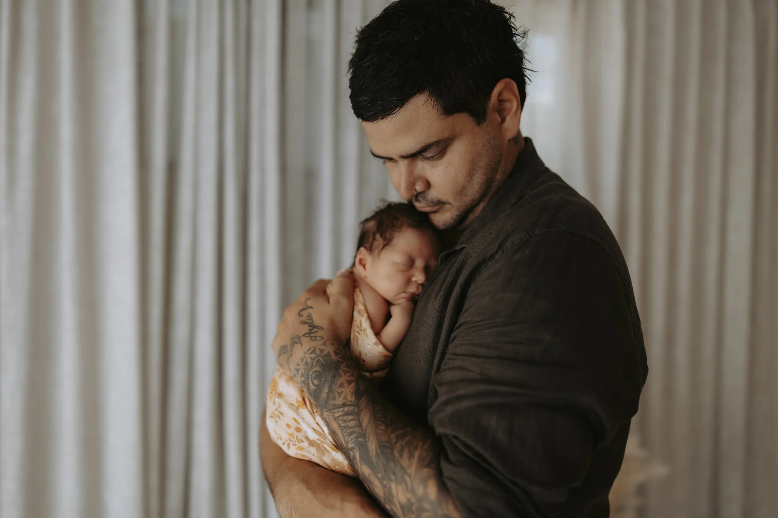 A man with tattoos on his arm holding a sleeping newborn baby close to his chest, with a neutral-colored curtain in the background.