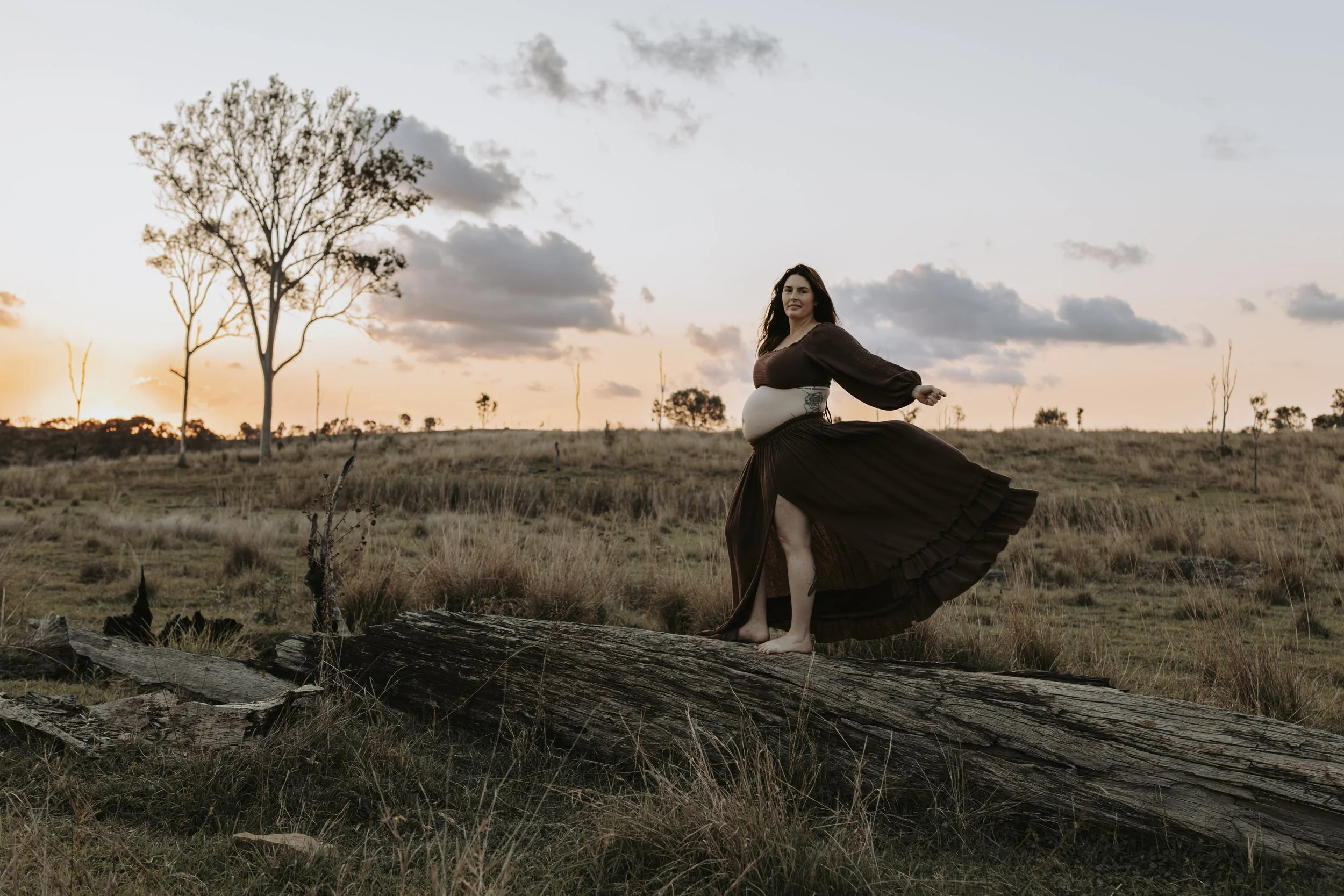 Pregnant woman standing barefoot on a fallen log in a grassy field at sunset, wearing a long dark dress with a high slit, with trees and a cloudy sky in the background.