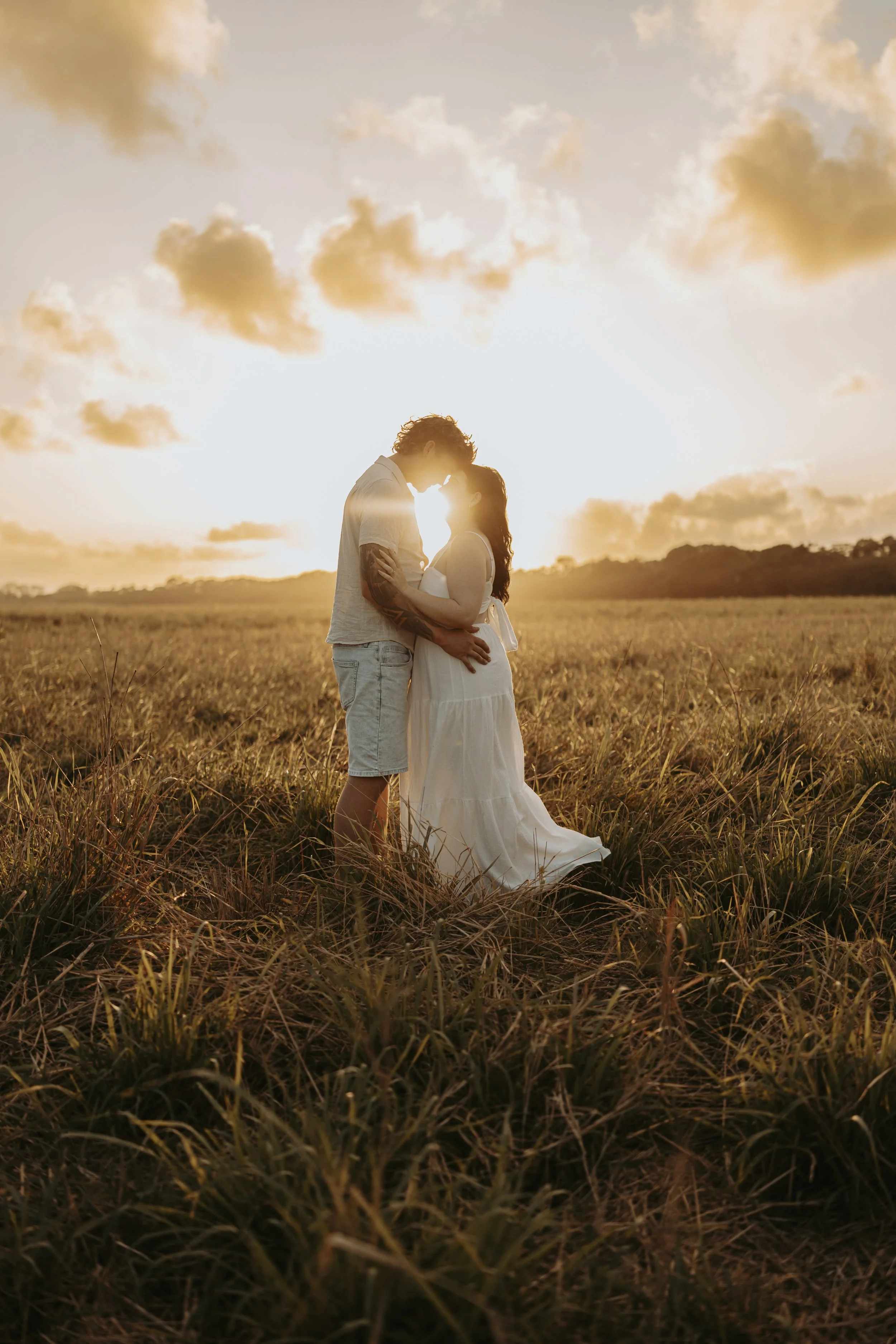 A couple standing in a field during sunset, touching foreheads, with the sun behind them creating a glow around their silhouettes.