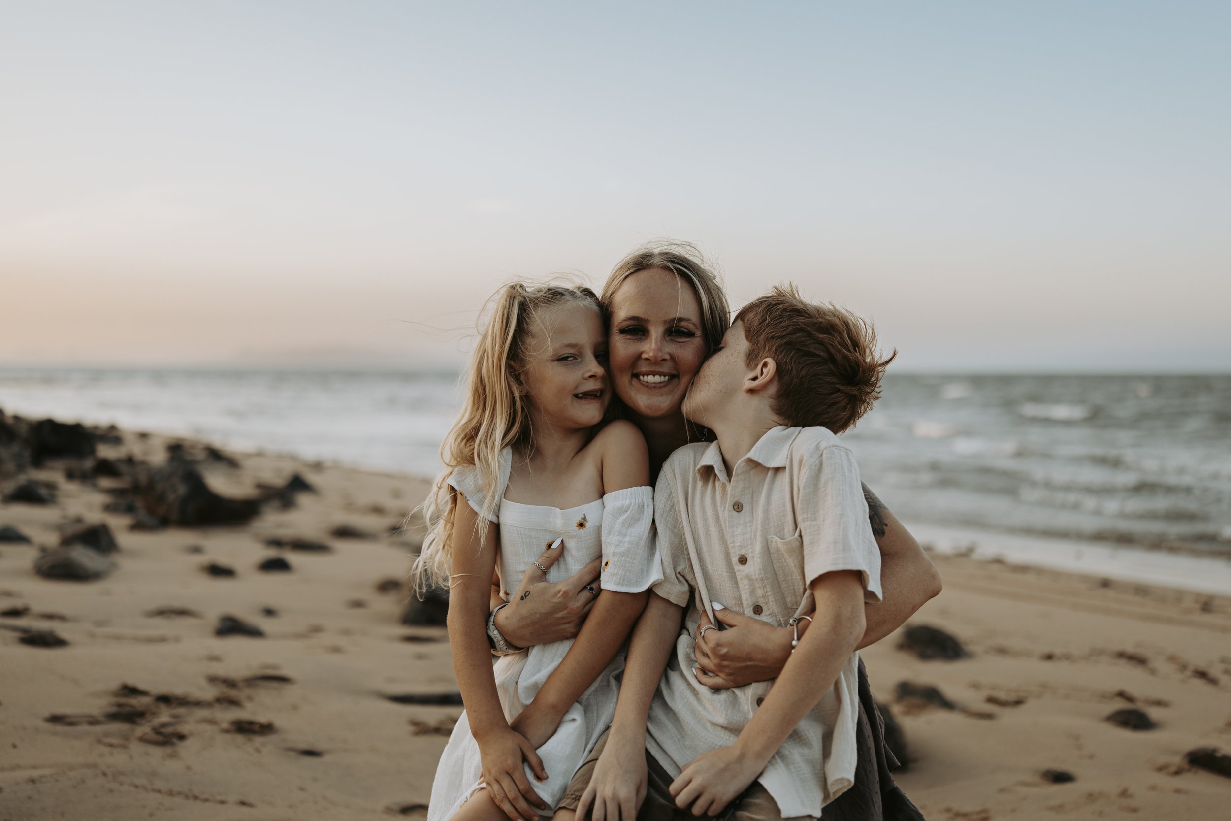 A woman with two children, a girl and a boy, on a beach during sunset. The woman and girl are smiling, and the boy is kissing the woman's cheek. The scene is warm and joyful with the ocean and rocks in the background.