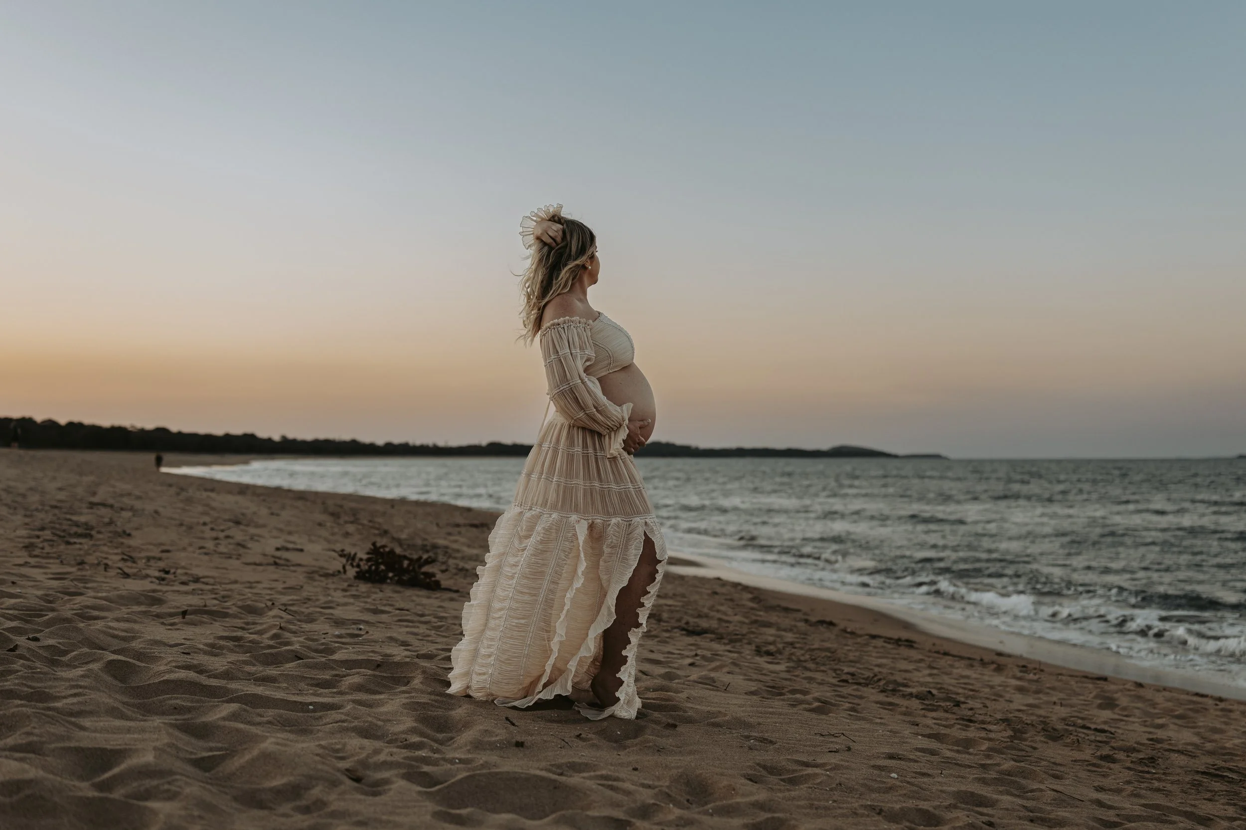 A pregnant woman in a beige dress standing on a sandy beach, looking at the ocean during sunset.