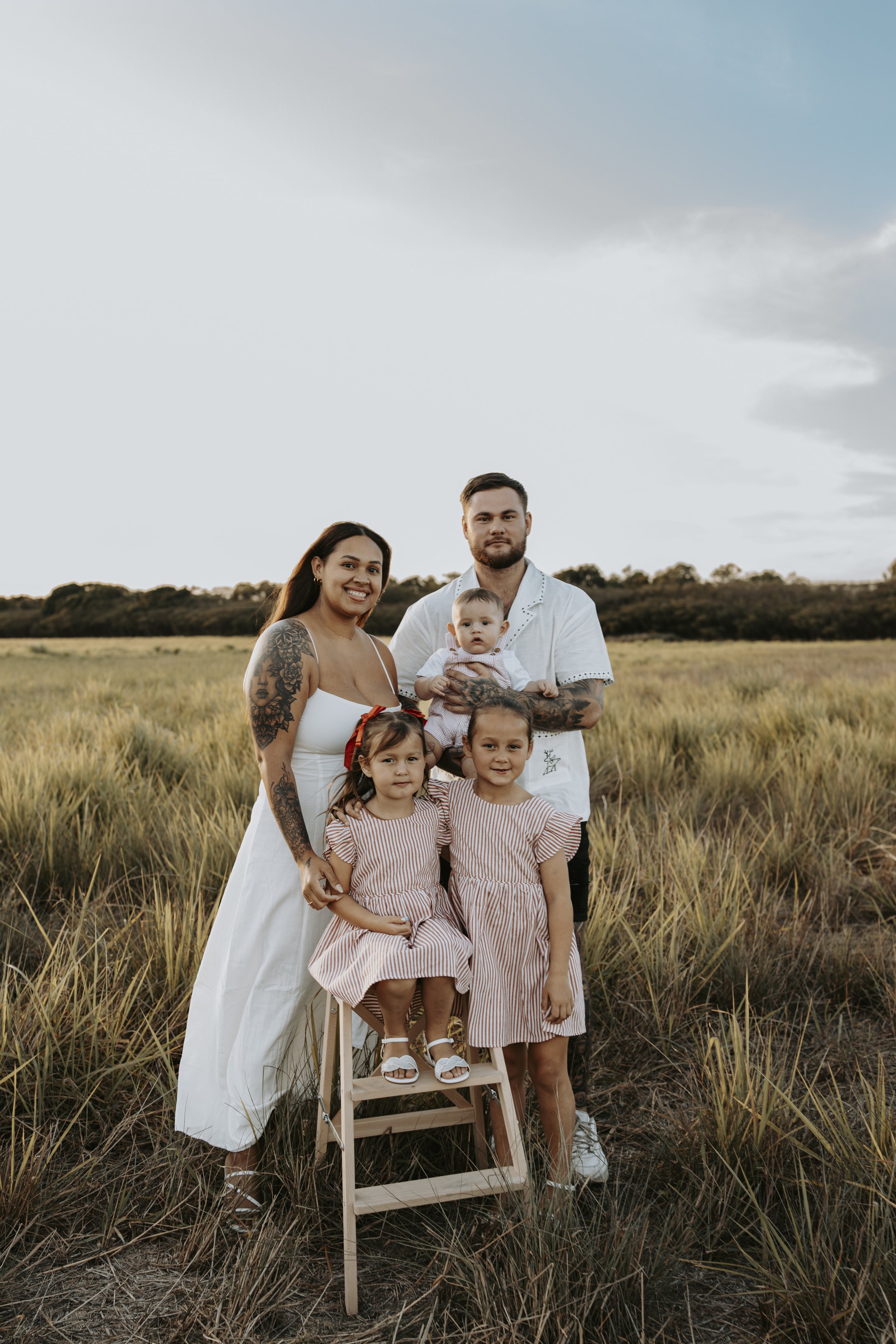 Family of five standing in a grassy field during sunset, smiling at the camera. The mother has tattoos and is wearing a white dress, while the father holds a baby. Two young girls in striped dresses sit on a small wooden step stool.