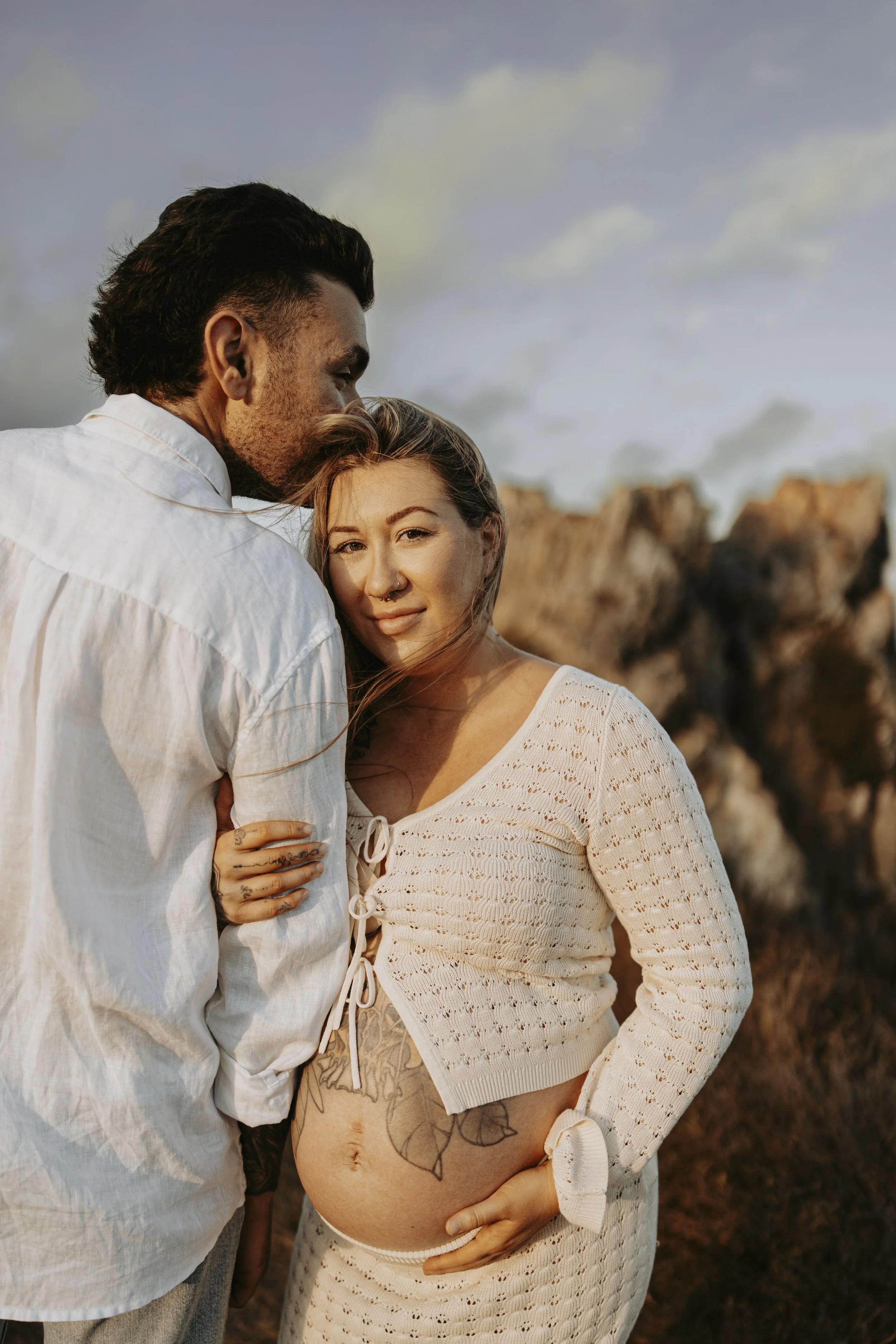 A pregnant woman and a man standing outside, with the man kissing the top of the woman's head. The woman has tattoos on her belly and wears a cream-colored knit top, while the man wears a white shirt. The background features a natural landscape with 