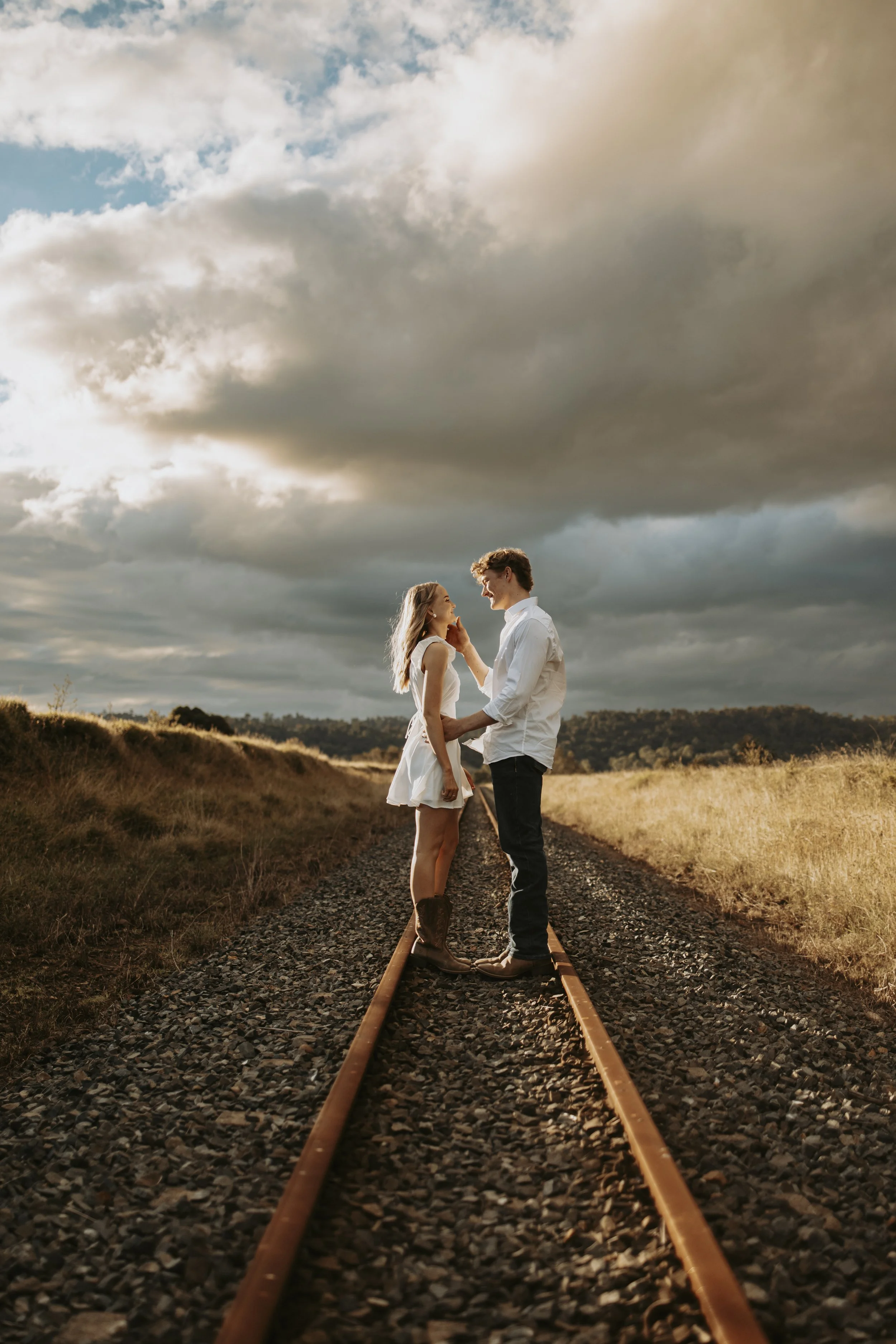 A couple standing on train tracks in an open field during sunset, with dramatic clouds overhead.