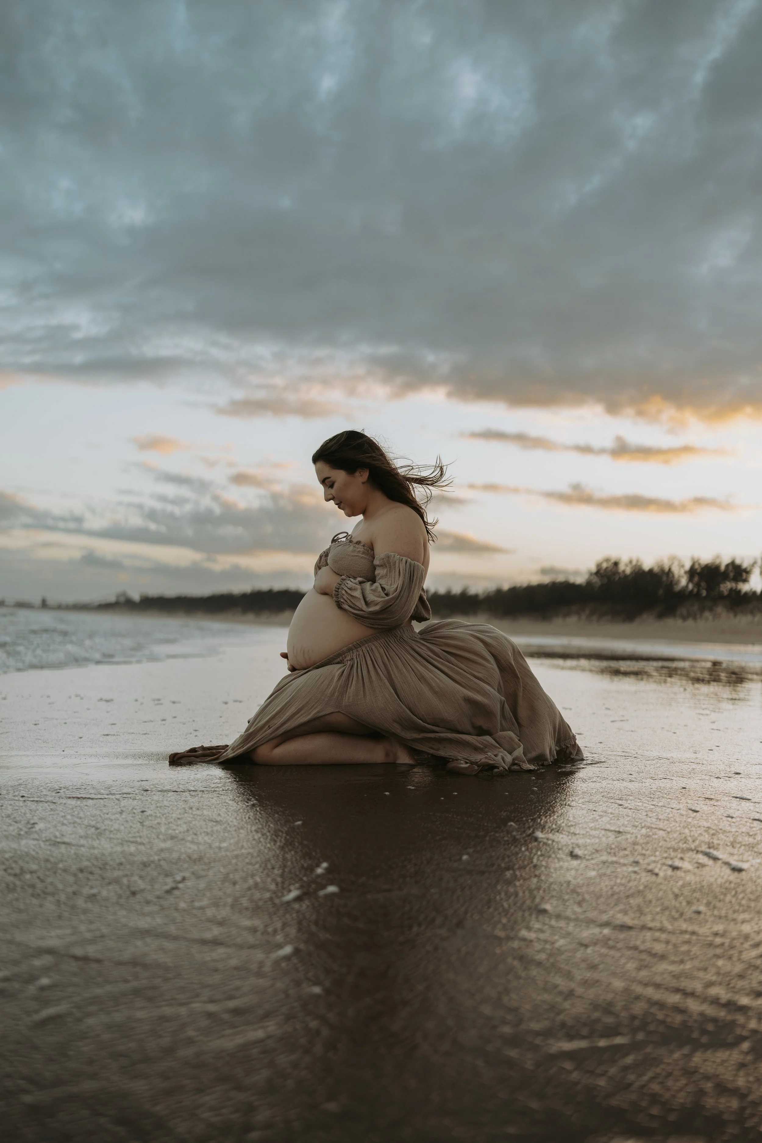 A pregnant woman kneeling on a beach at sunset with her hands on her belly, wearing a flowing beige dress, with her hair blowing in the wind.