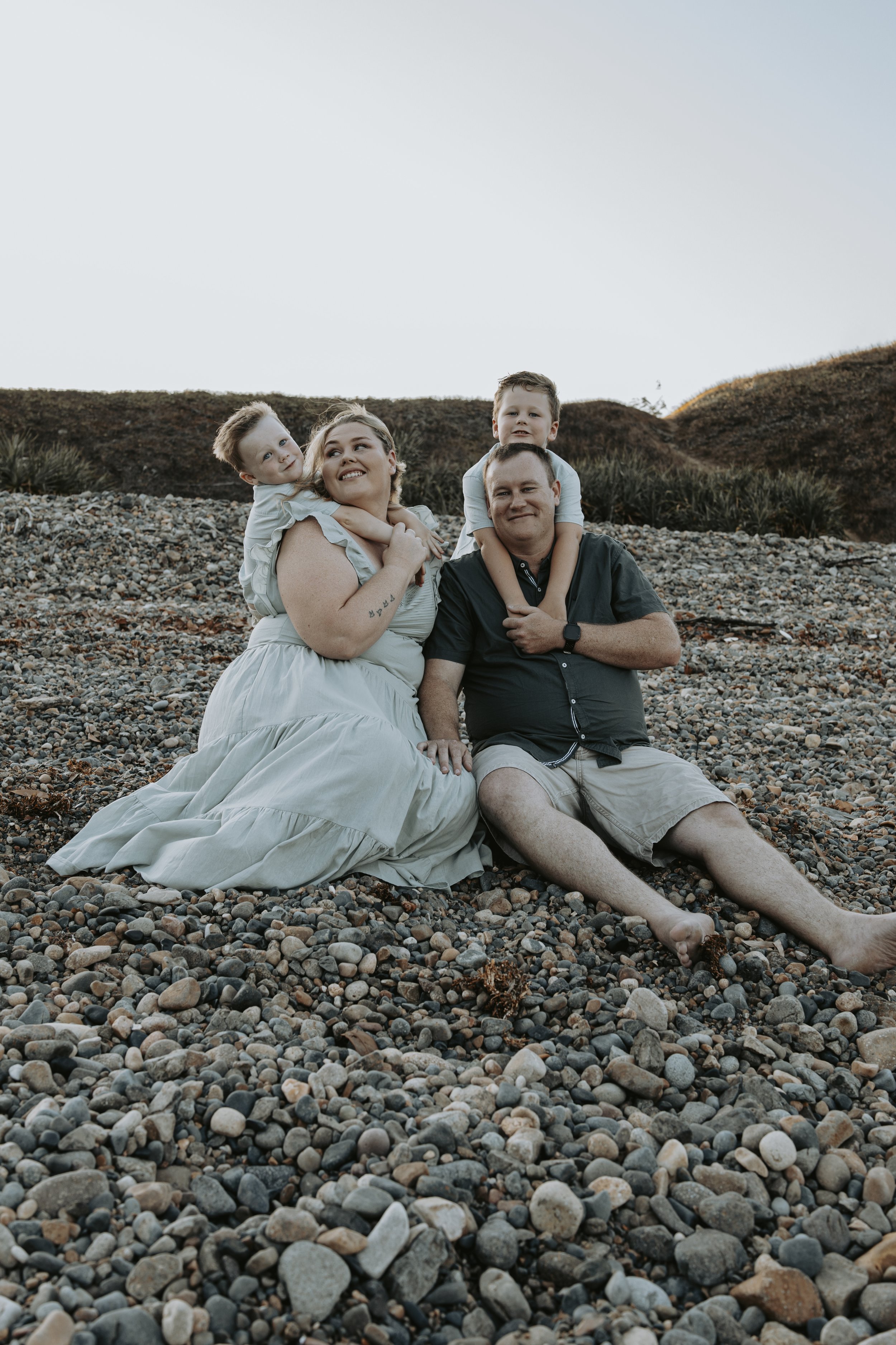 Family of four (mother, father, and two young boys) sitting on a rocky beach with hills in the background, enjoying a moment together.