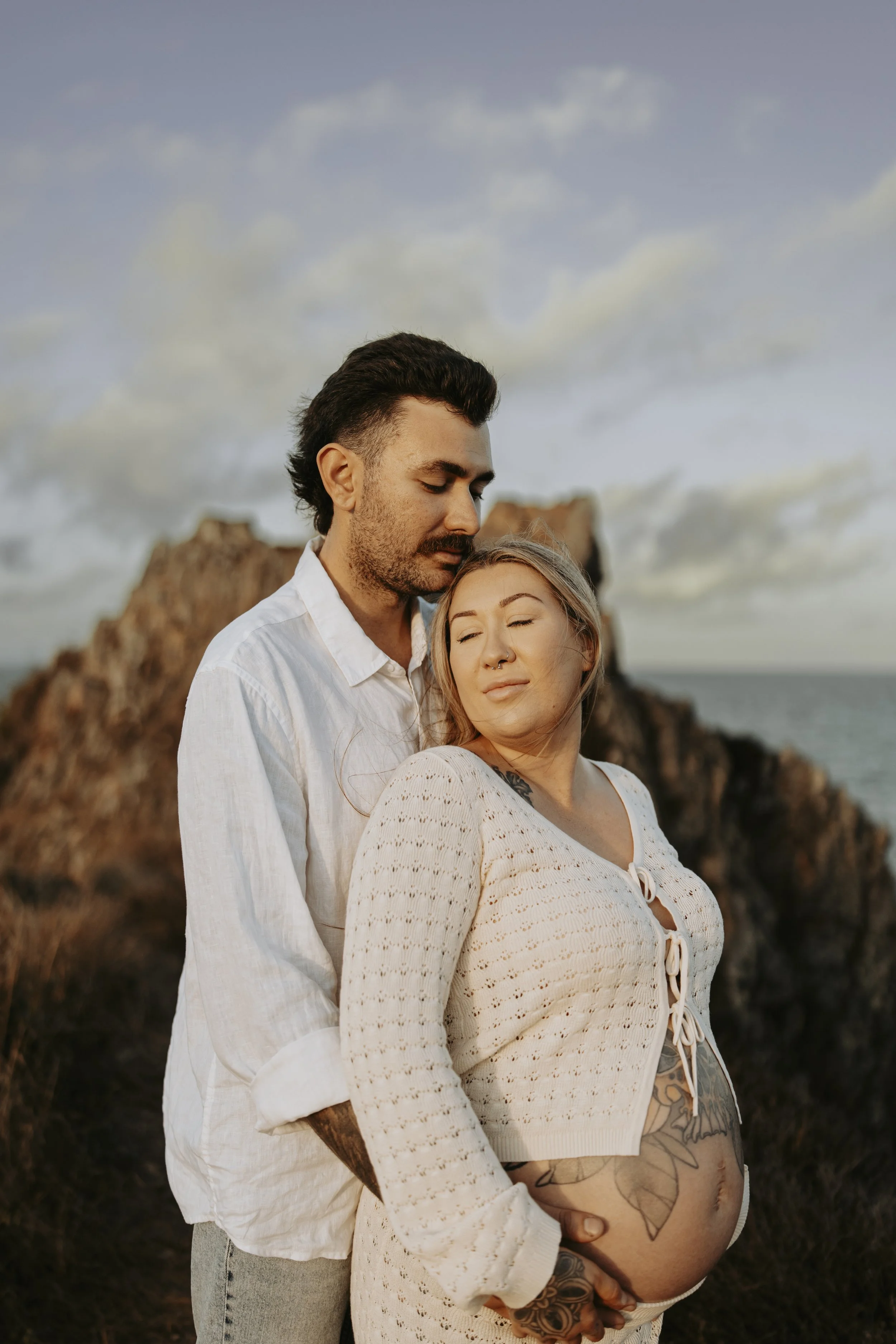 A couple stands close together outdoors, with the man embracing the pregnant woman from behind. They are in a natural setting with rocks and a cloudy sky in the background. The woman has tattoos and is wearing a cream cardigan, and the man has dark h