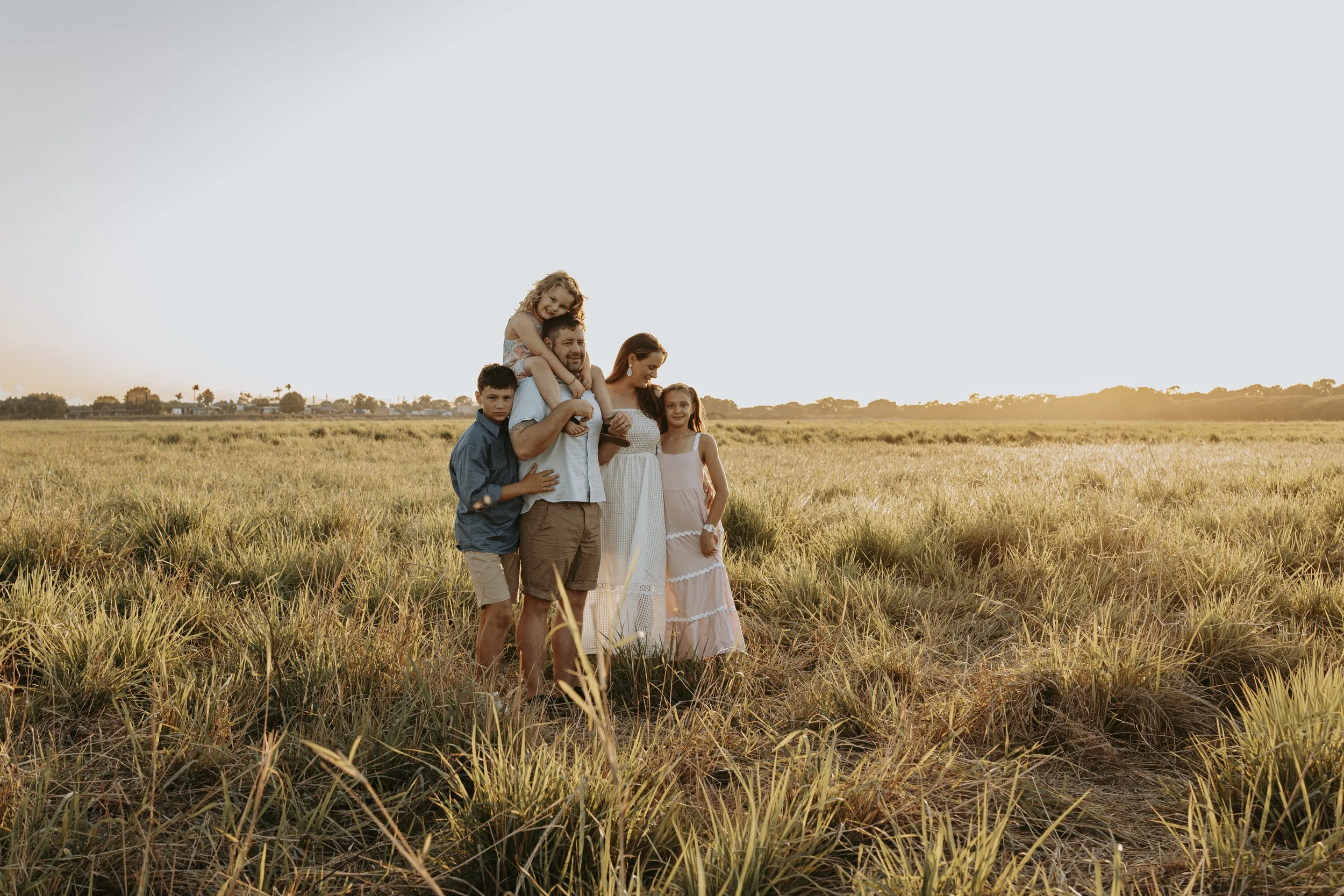 Family of six standing in a field at sunset, with the father carrying a young girl on his shoulders and others gathered around him.