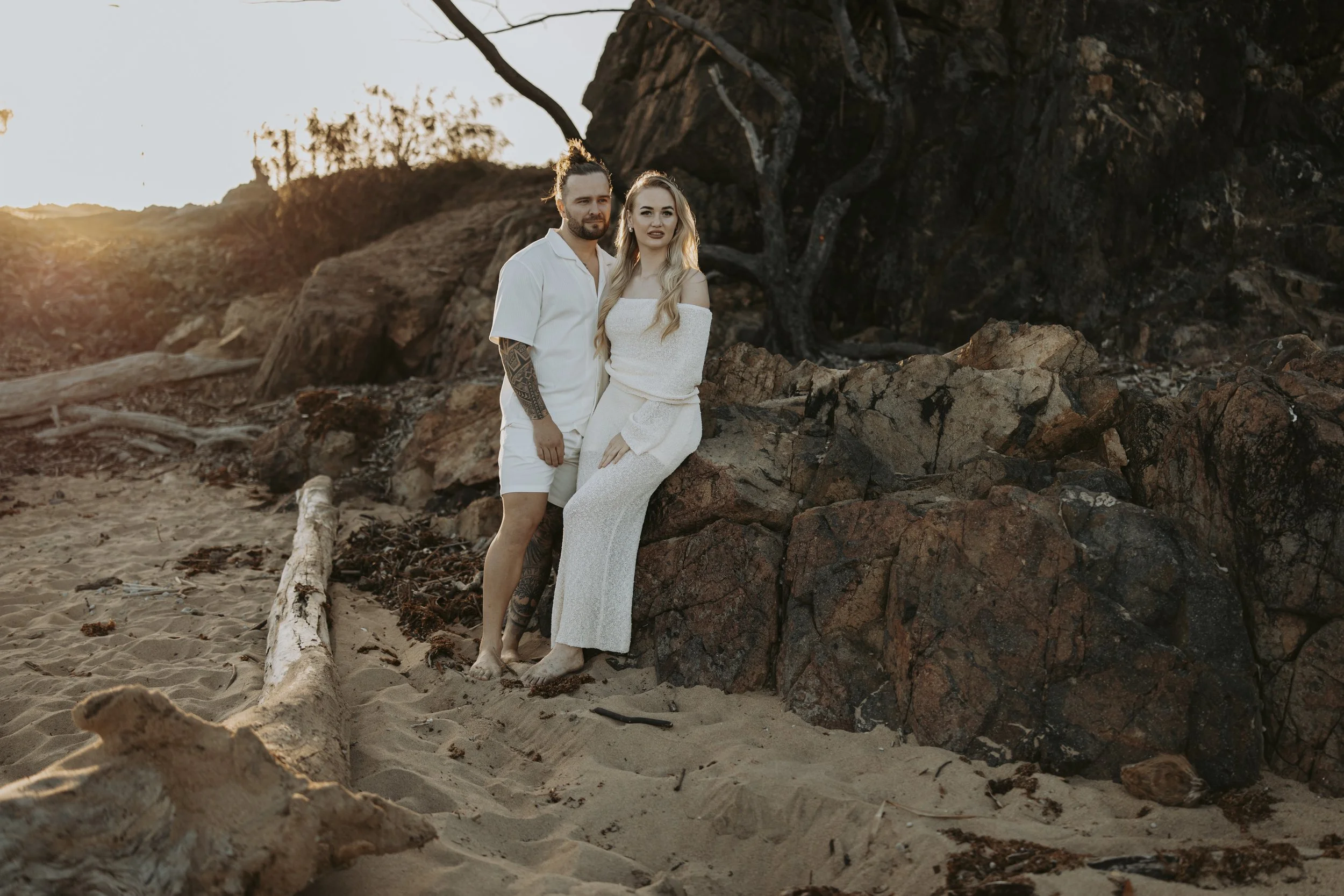 A couple standing barefoot on a sandy beach near rocks at sunset. The man has tattoos on his arm and is wearing a white shirt and shorts, while the woman is in a white off-shoulder dress. They are positioned close together, with the man holding the w