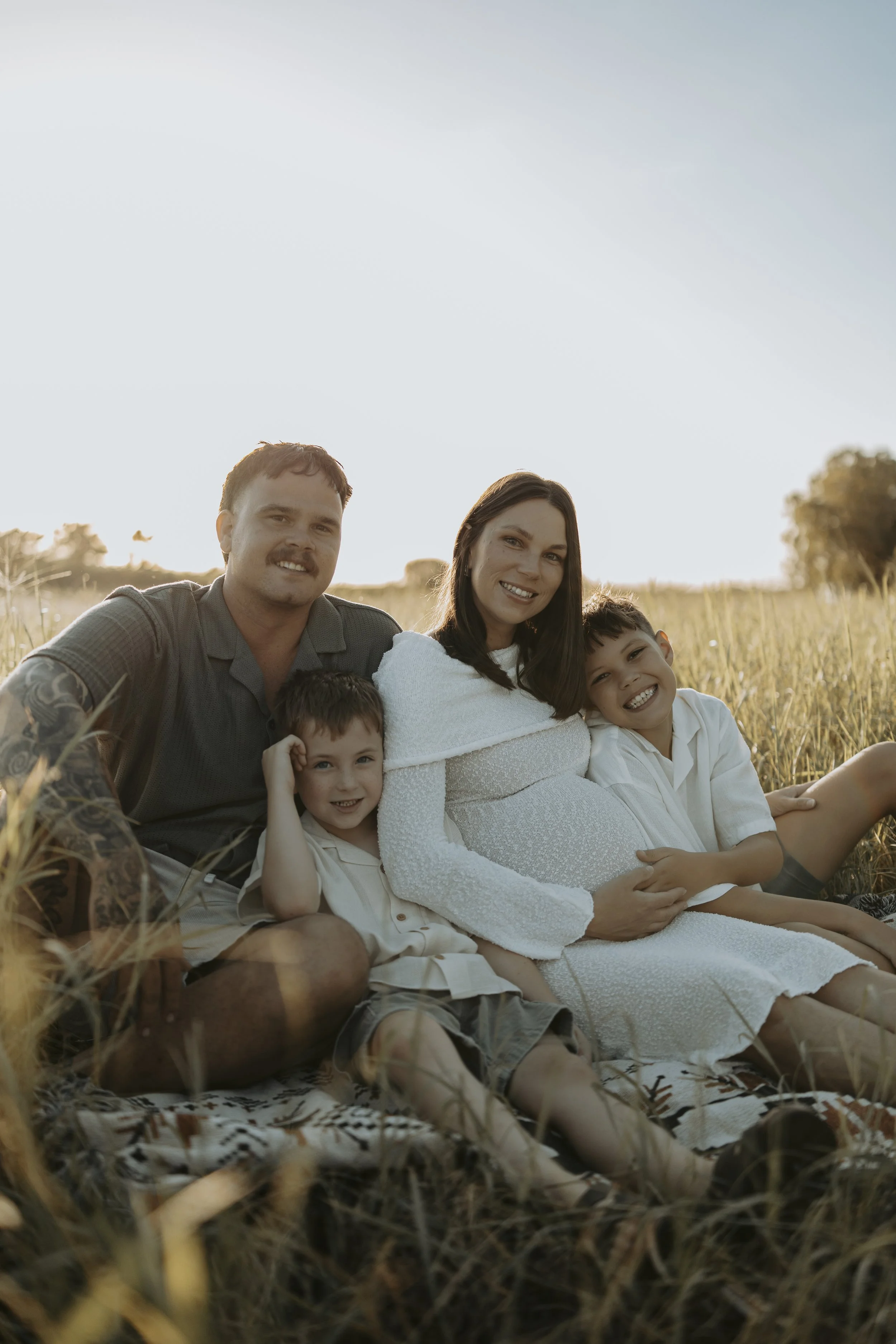 A family of five sitting on a blanket in a field during sunset, with the mother visibly pregnant, smiling and enjoying each other's company.