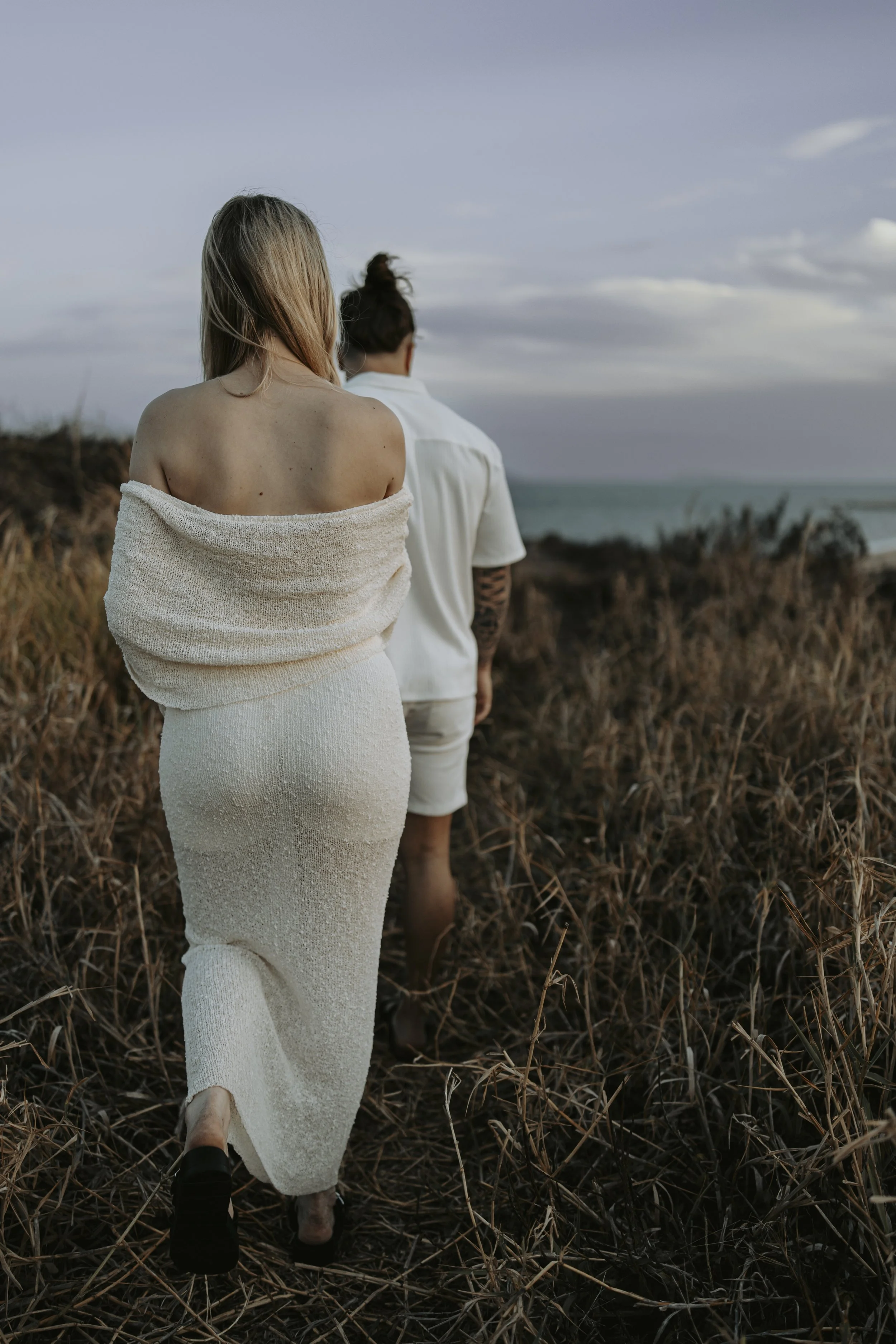 Two women walking through dry grass towards the beach, with ocean and cloudy sky in the background.