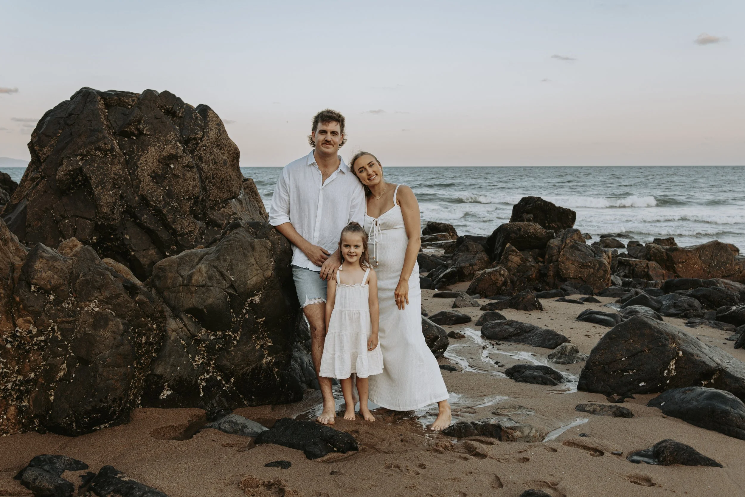 A family of three posing on a beach with rocks and ocean in the background during sunset. The man is standing with his arm around the woman, who is pregnant, and the young girl is standing in front of them. All are dressed in white or light-colored c
