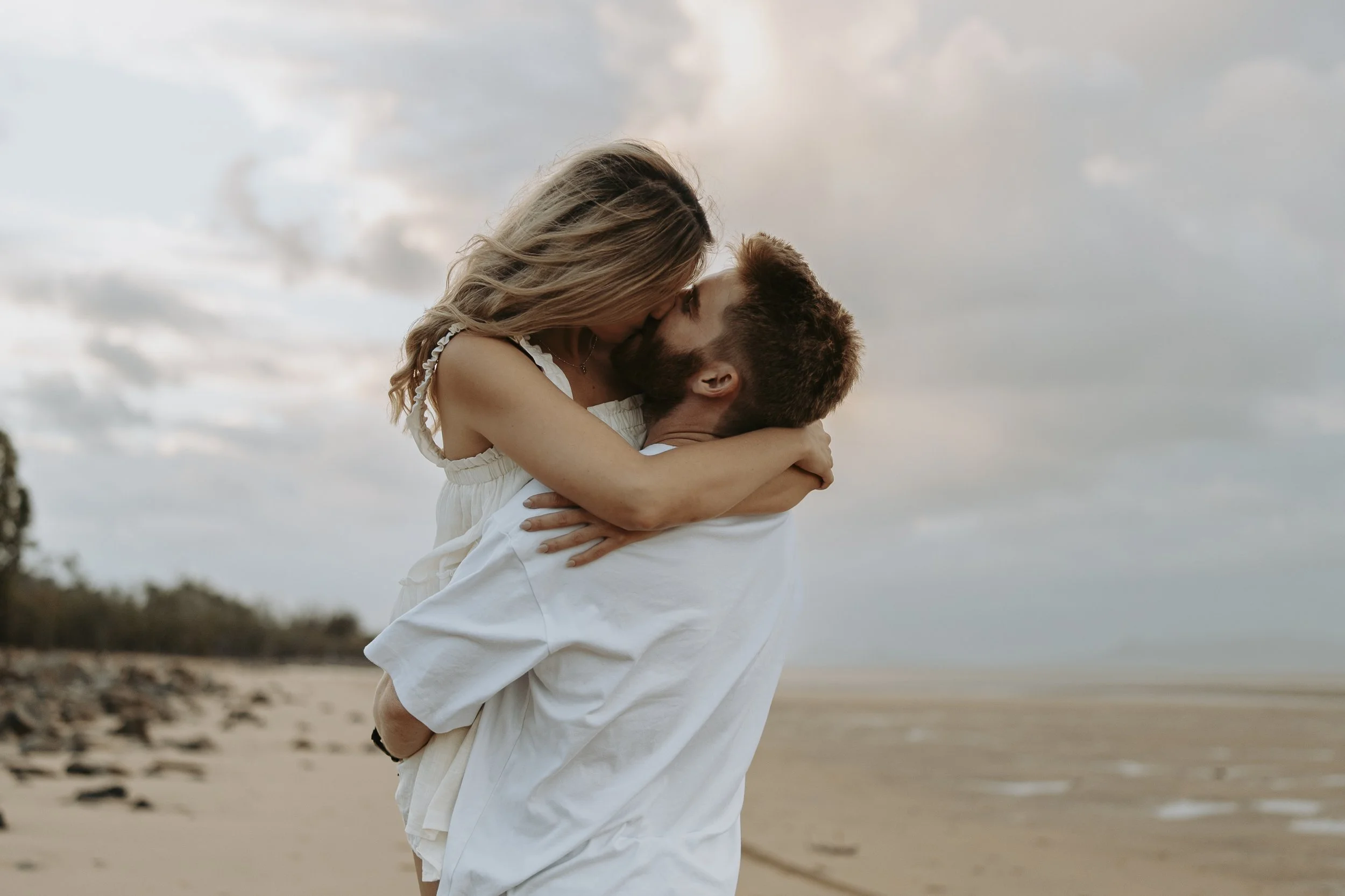 A couple embracing and kissing on a beach during sunset or dusk.