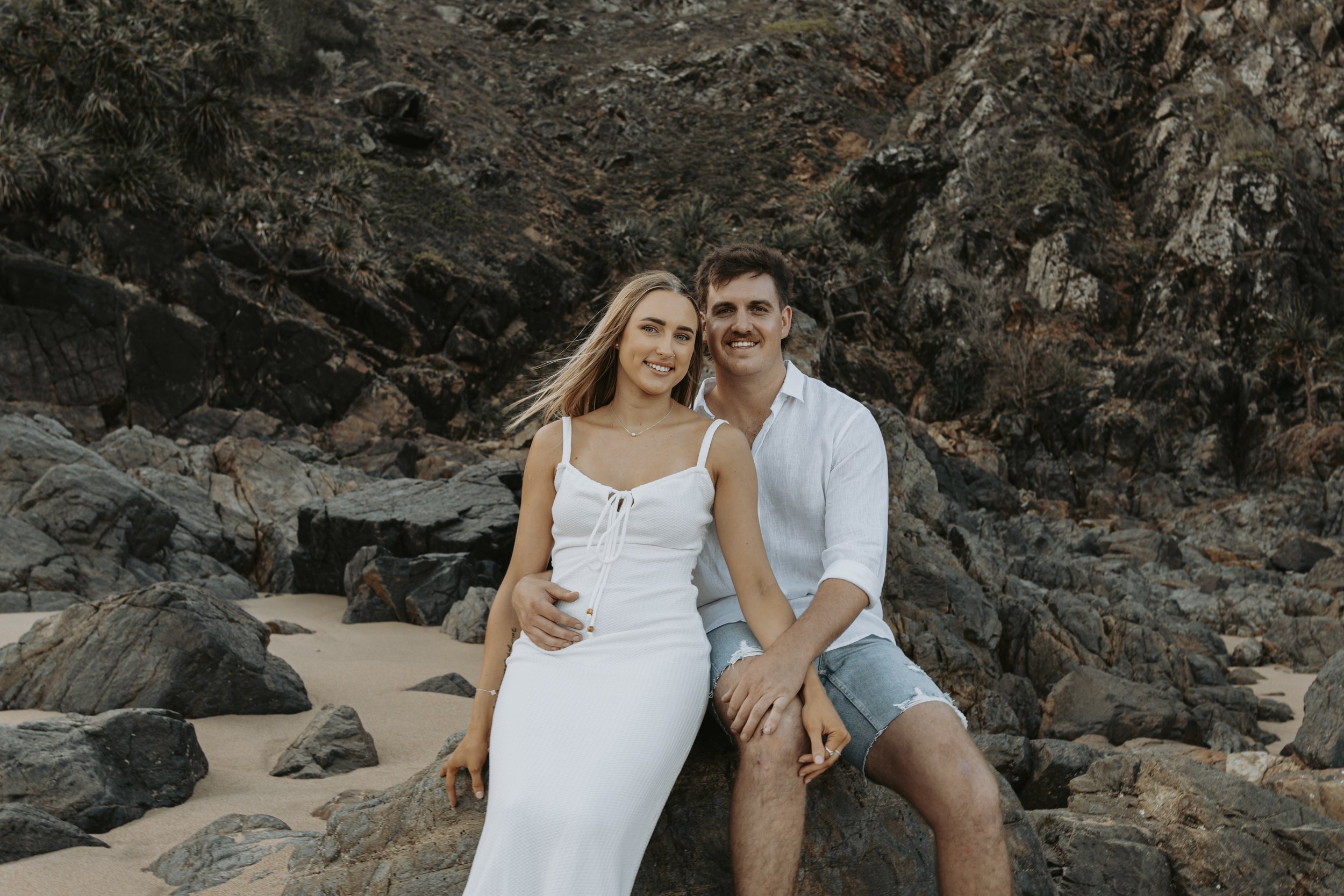 A young couple sitting on rocks on a beach with a rocky hillside behind them. The woman is wearing a white dress, and the man is wearing a white shirt with rolled-up sleeves and denim shorts. They are smiling and looking at the camera.