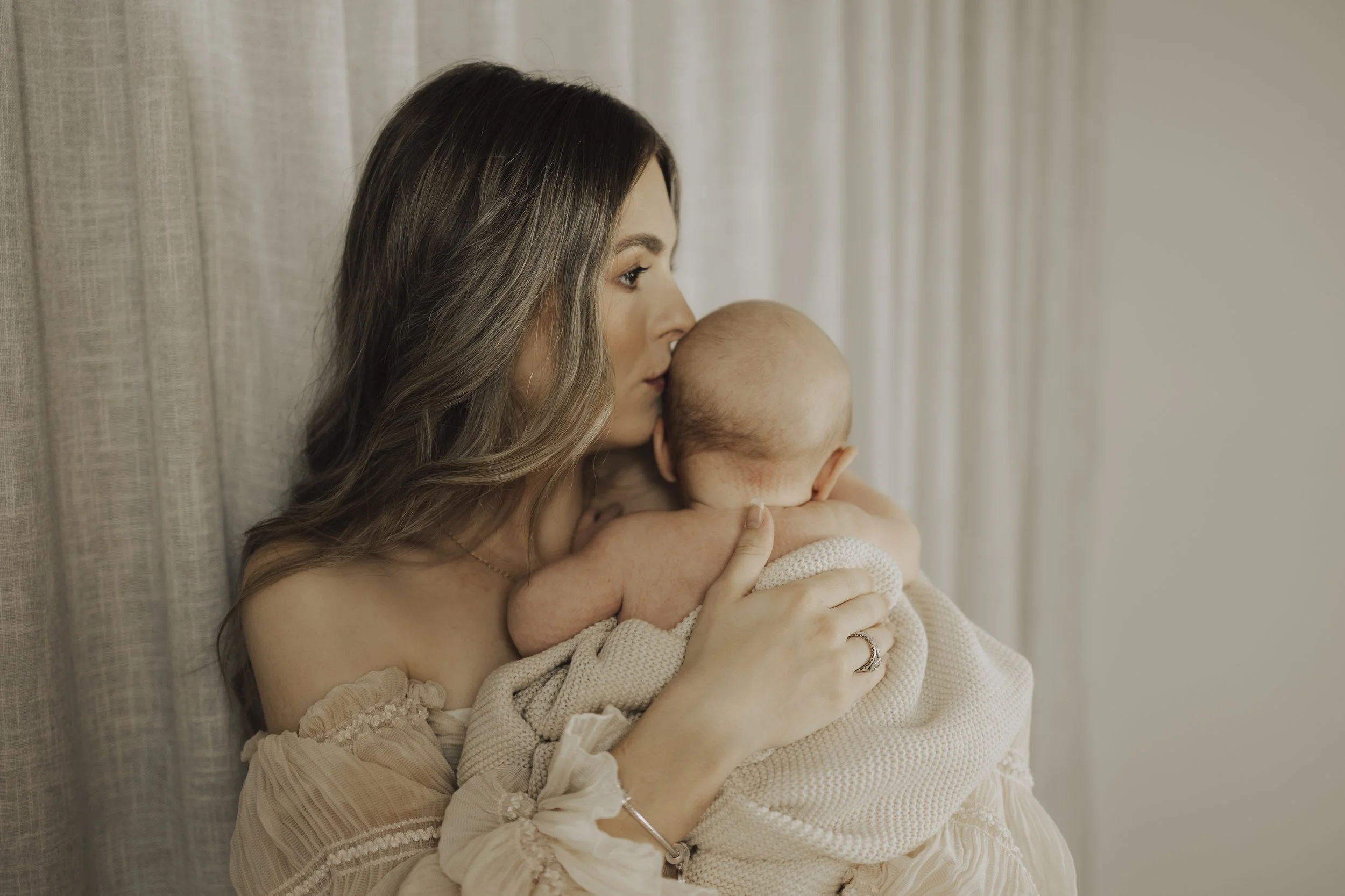 Woman holding a bald baby, gently touching their back, near a curtain.