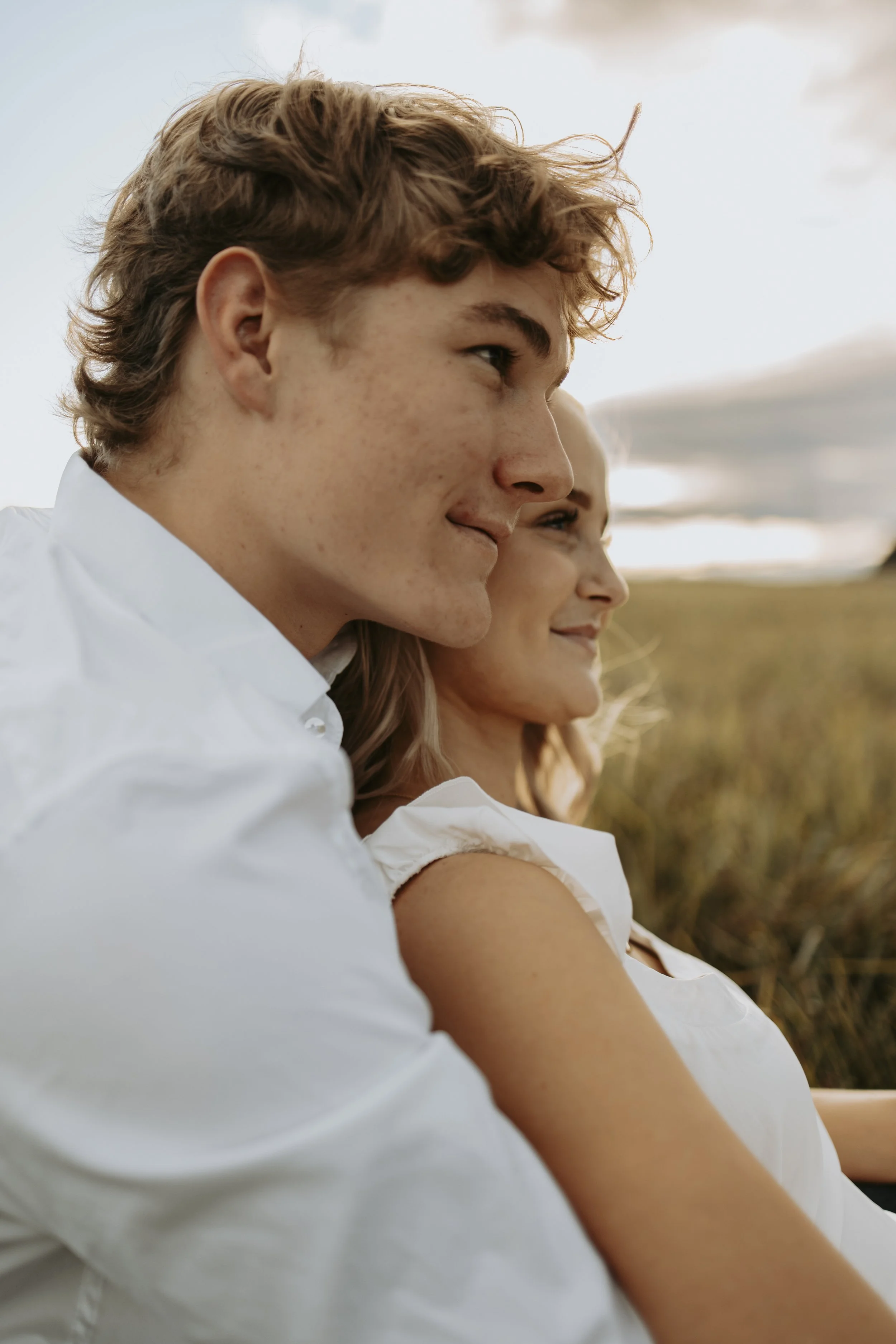 A young man and woman sitting outdoors in a field during sunset, smiling and looking to the right.