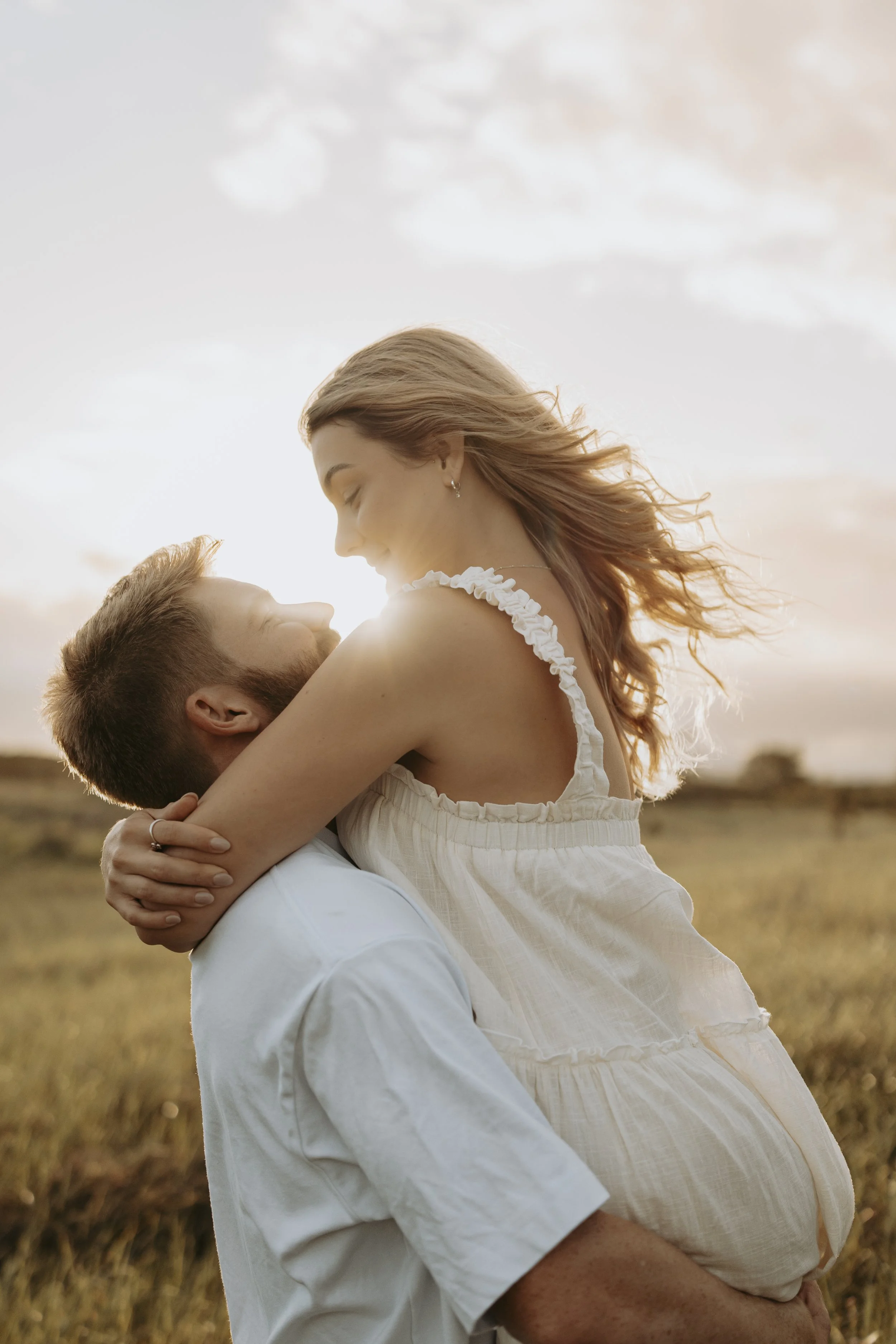 A couple embraces outdoors at sunset, with the woman sitting on the man's shoulders, smiling and looking into each other's eyes.
