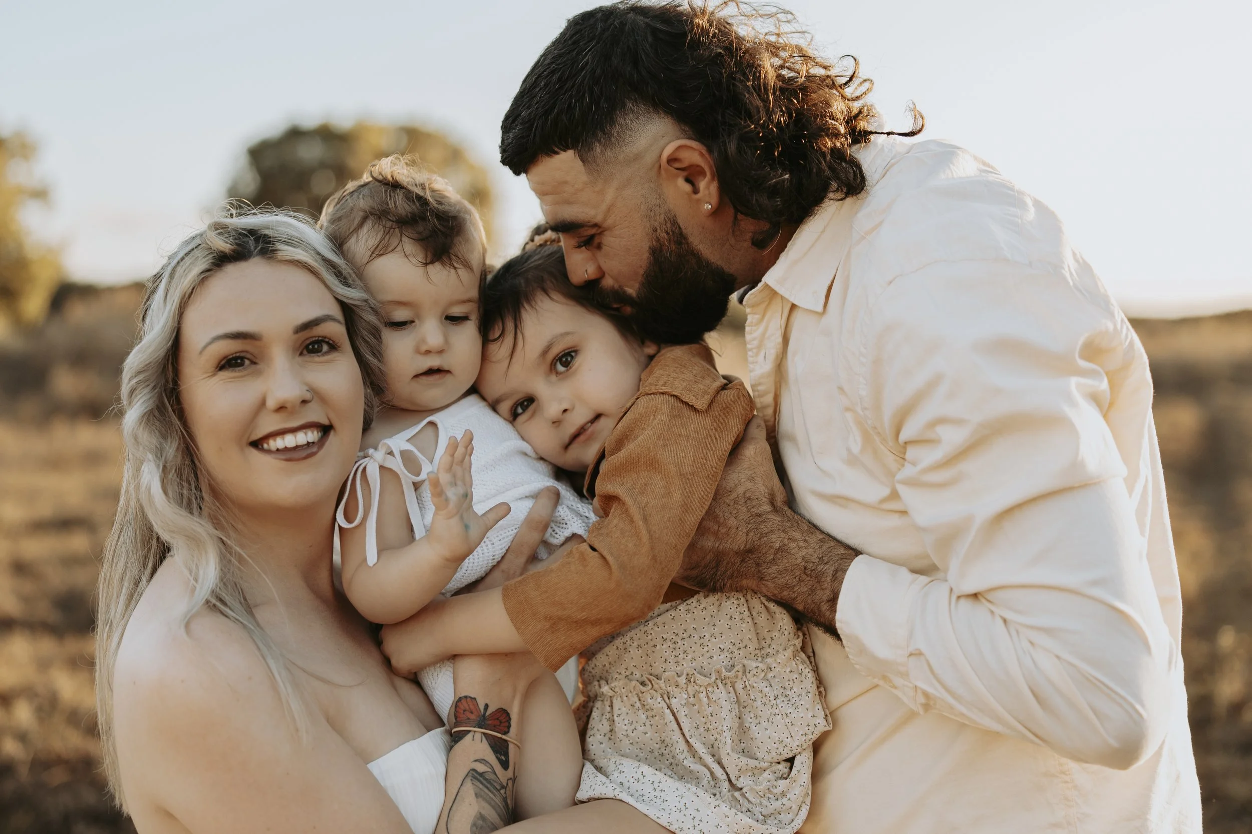 Family of four, a woman with platinum blonde hair, a man with a beard and long hair, and two children, sharing a joyful moment outdoors during sunset.