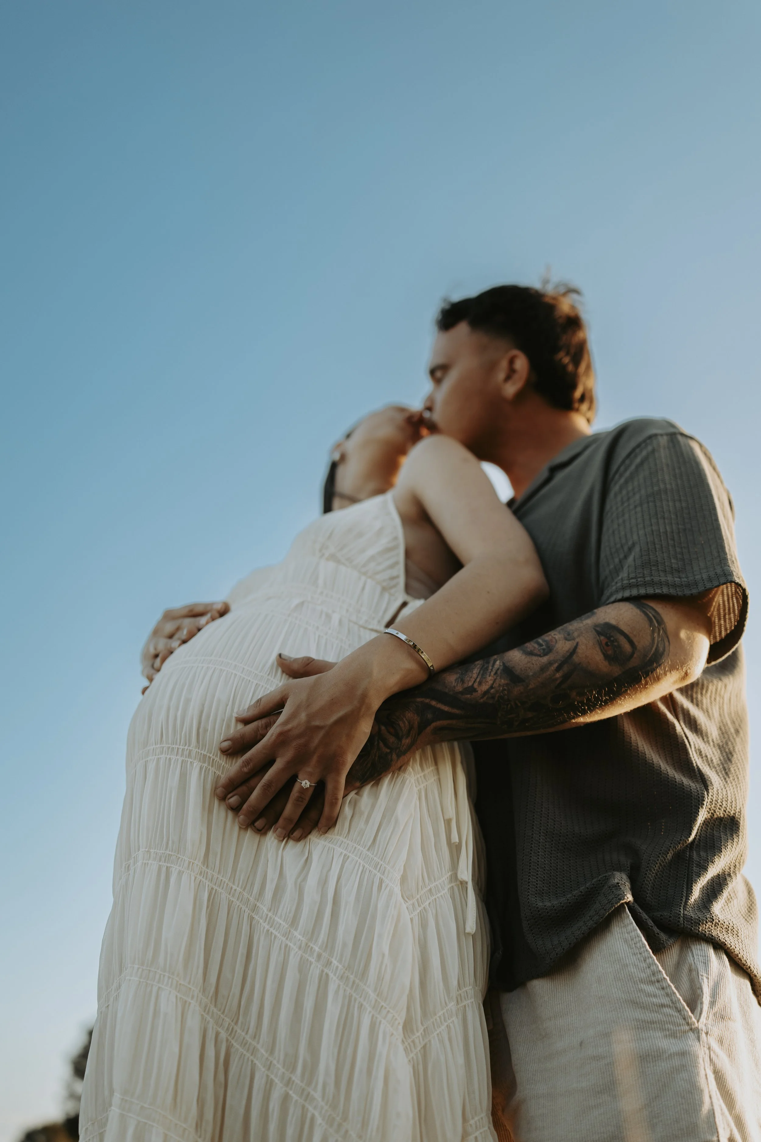 A couple kissing outdoors during sunset, with the woman wearing a white dress and the man wearing a gray shirt.