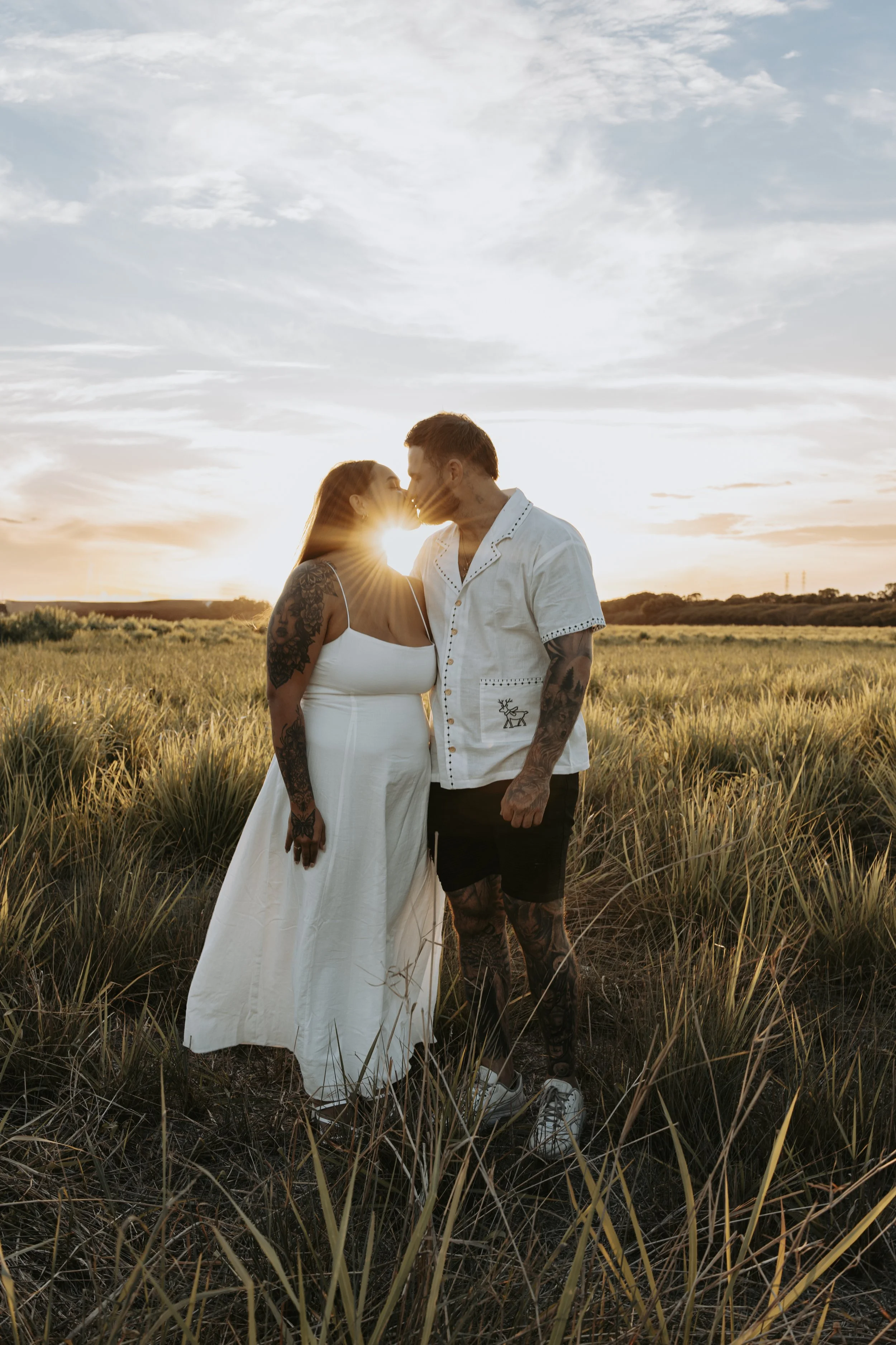 A couple leaning in for a kiss in a grassy field at sunset, with the sun partially obscured by their heads.