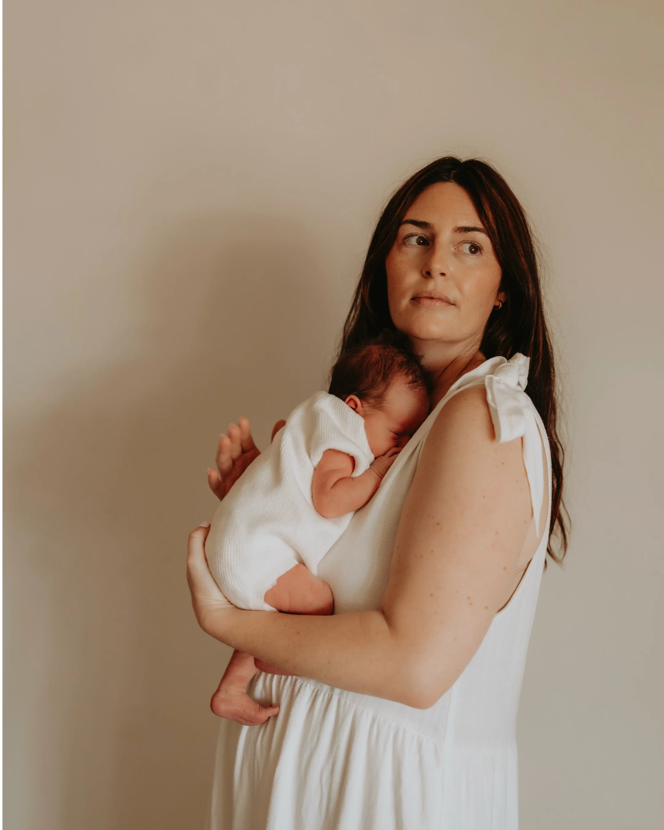 Woman holding a sleeping baby on her shoulder, standing against a plain wall.