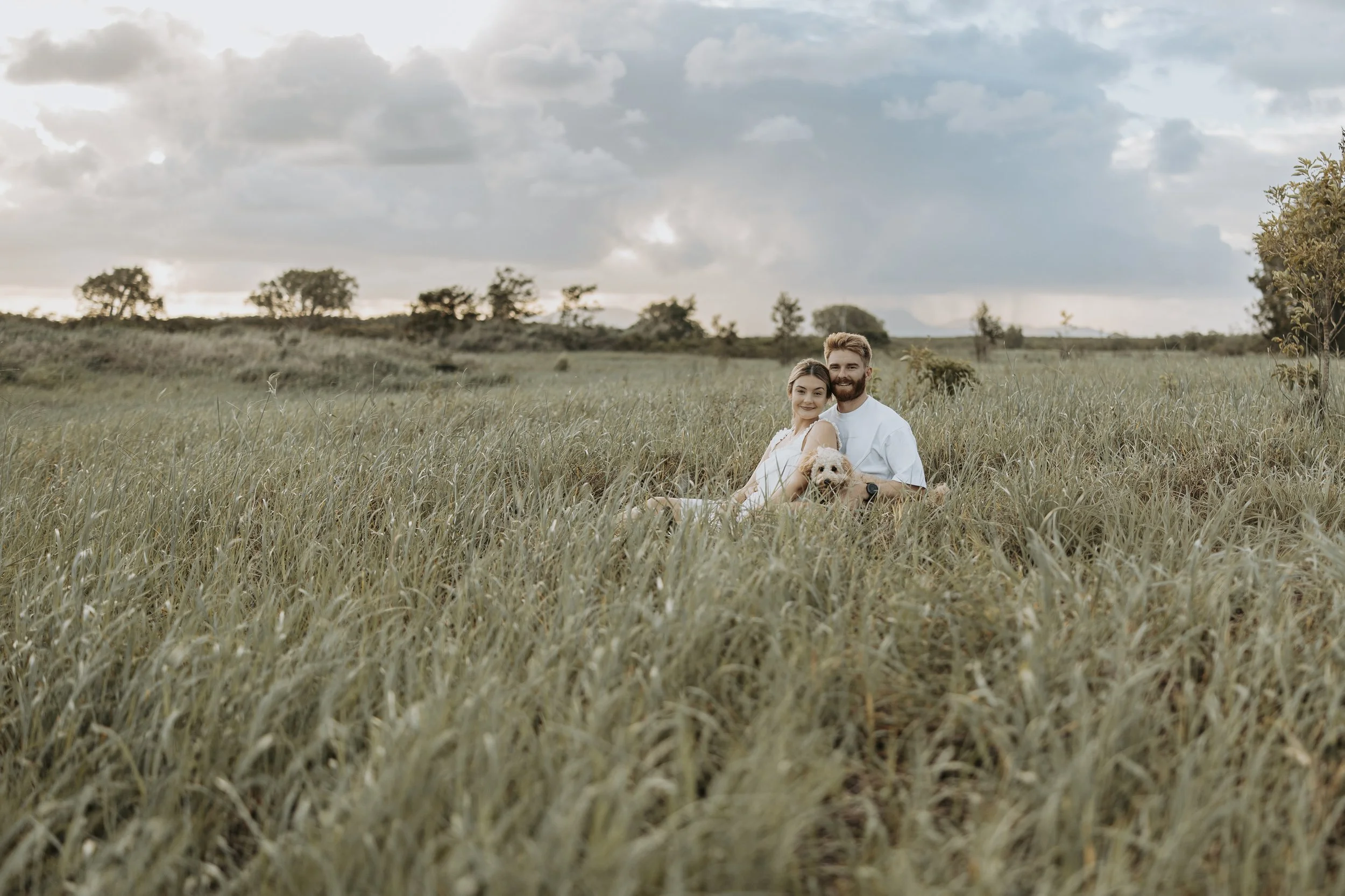 A couple sitting in a grassy field with their dog, under a partly cloudy sky, during sunset.