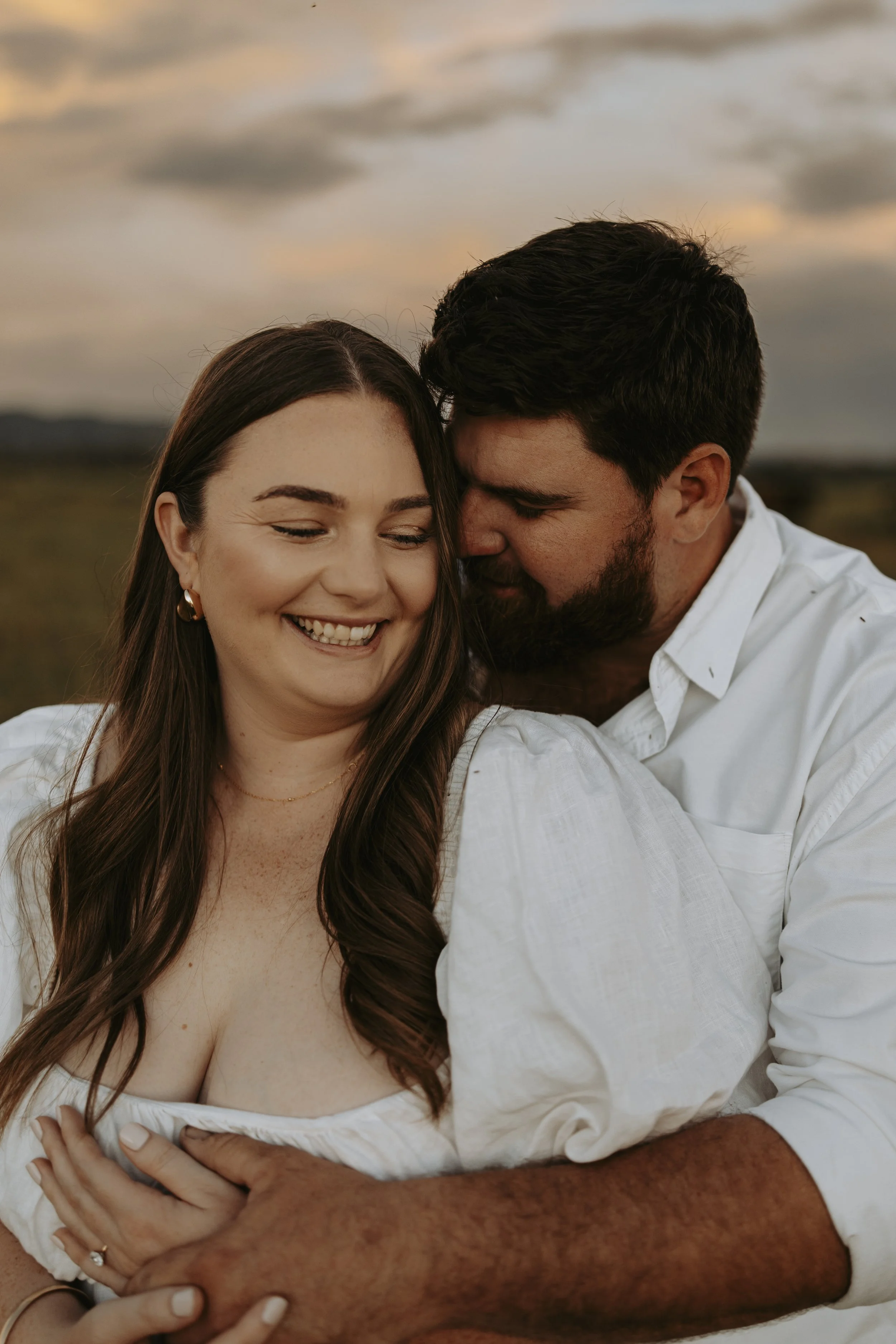 A smiling woman and a bearded man embracing outdoors during sunset, with a cloudy sky in the background.