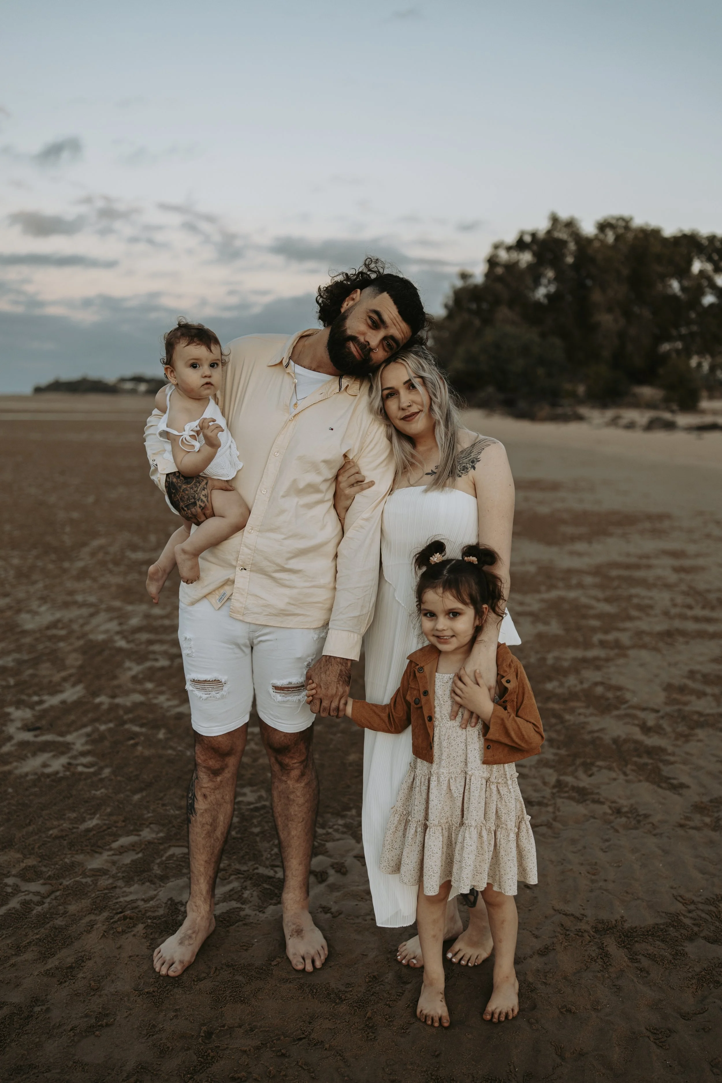 A family of four standing barefoot on a sandy beach during sunset, holding hands and embracing each other.
