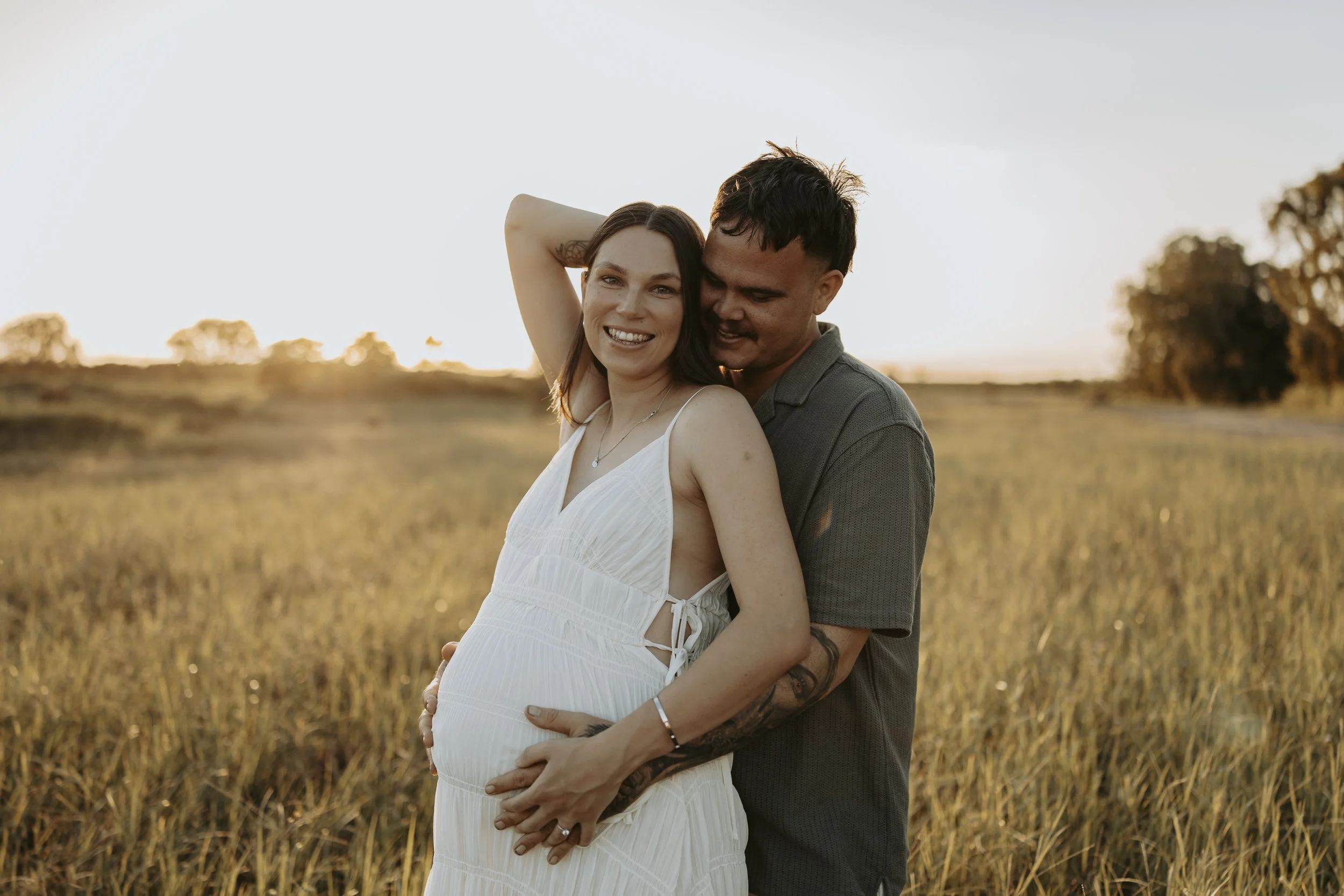 A pregnant woman in a white dress and a man in a gray shirt standing in a field at sunset, smiling and embracing each other.