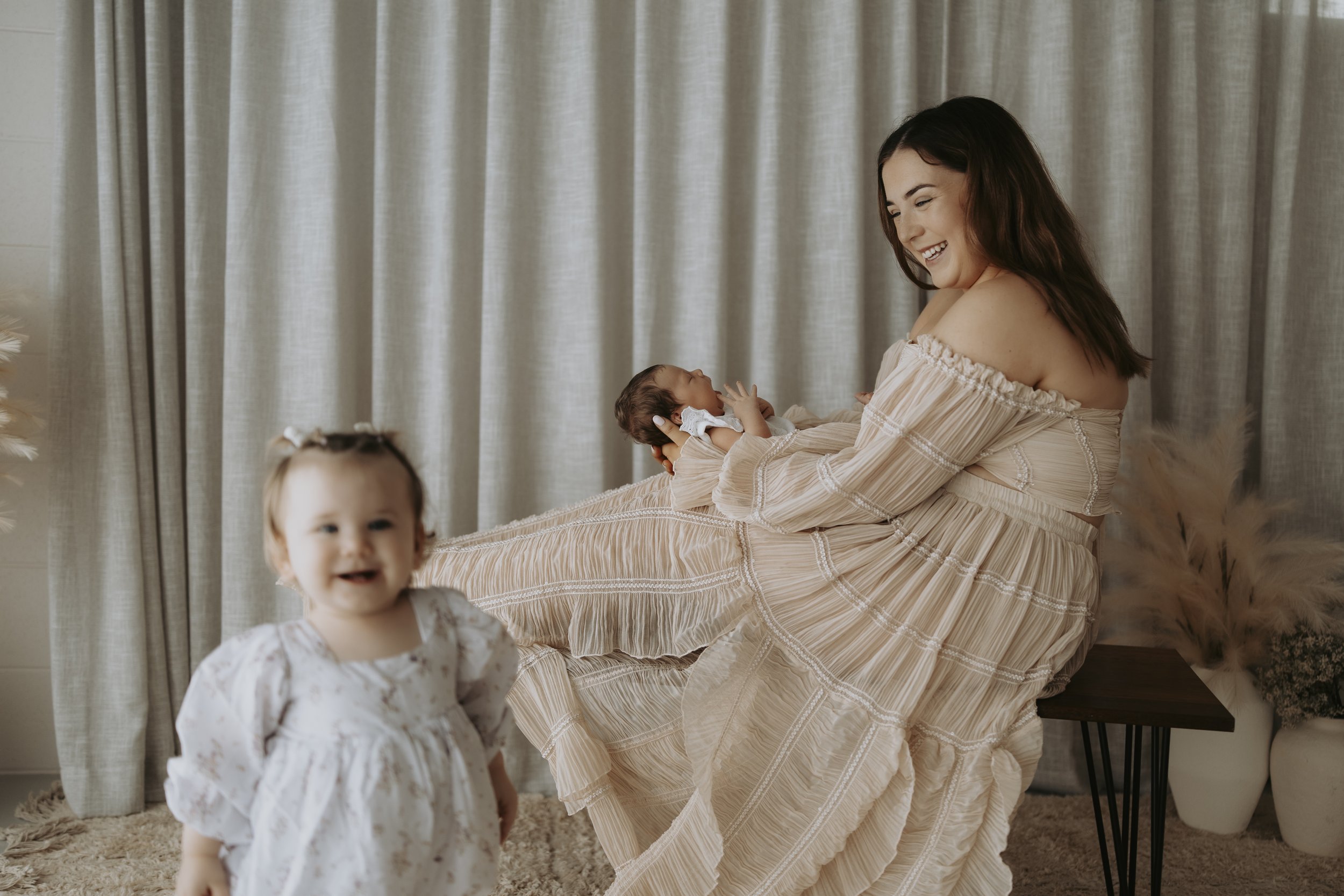 A woman is sitting on a bench holding a newborn baby while smiling at her. A toddler girl is standing in front of them, smiling at the camera. The setting appears to be indoors with neutral-colored curtains in the background.