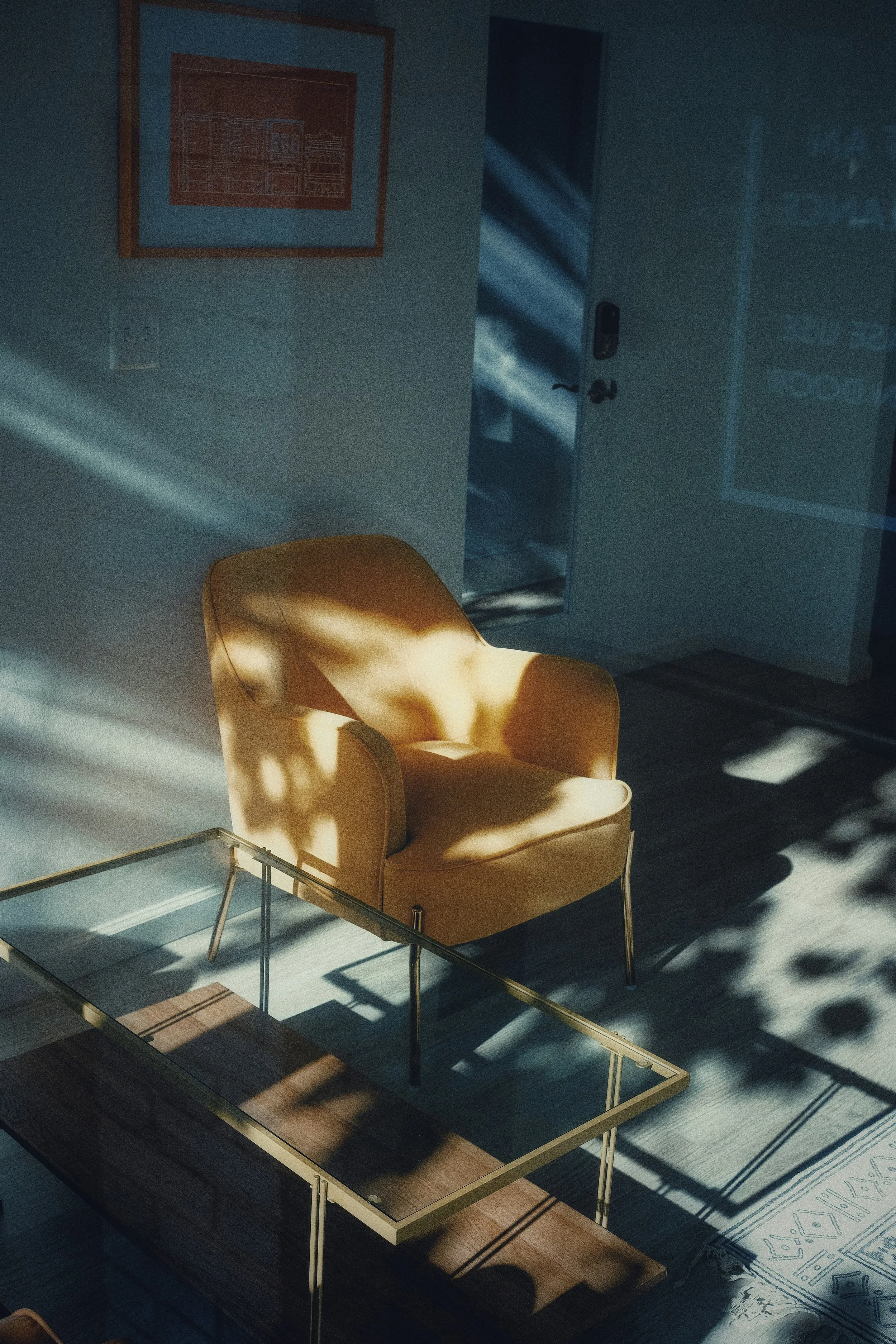 An empty orange armchair in a sunlit room with shadows on the walls and floor, a glass-top coffee table, and a framed artwork on the wall.